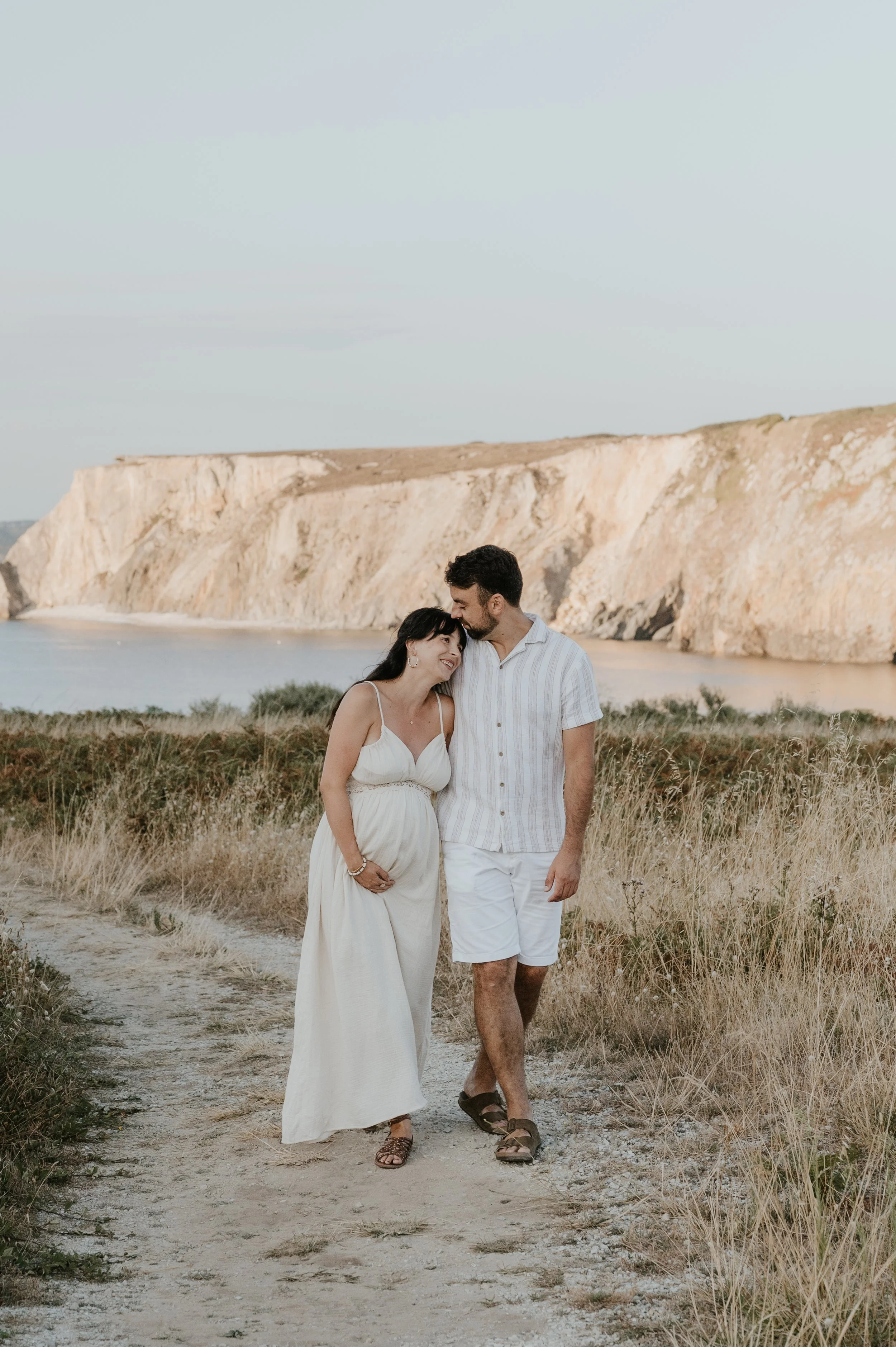 Un couple marche sur un sentier à la plage avec des falaises en arrière-plan lors du coucher de soleil, ils se tiennent la main et semblent heureux.