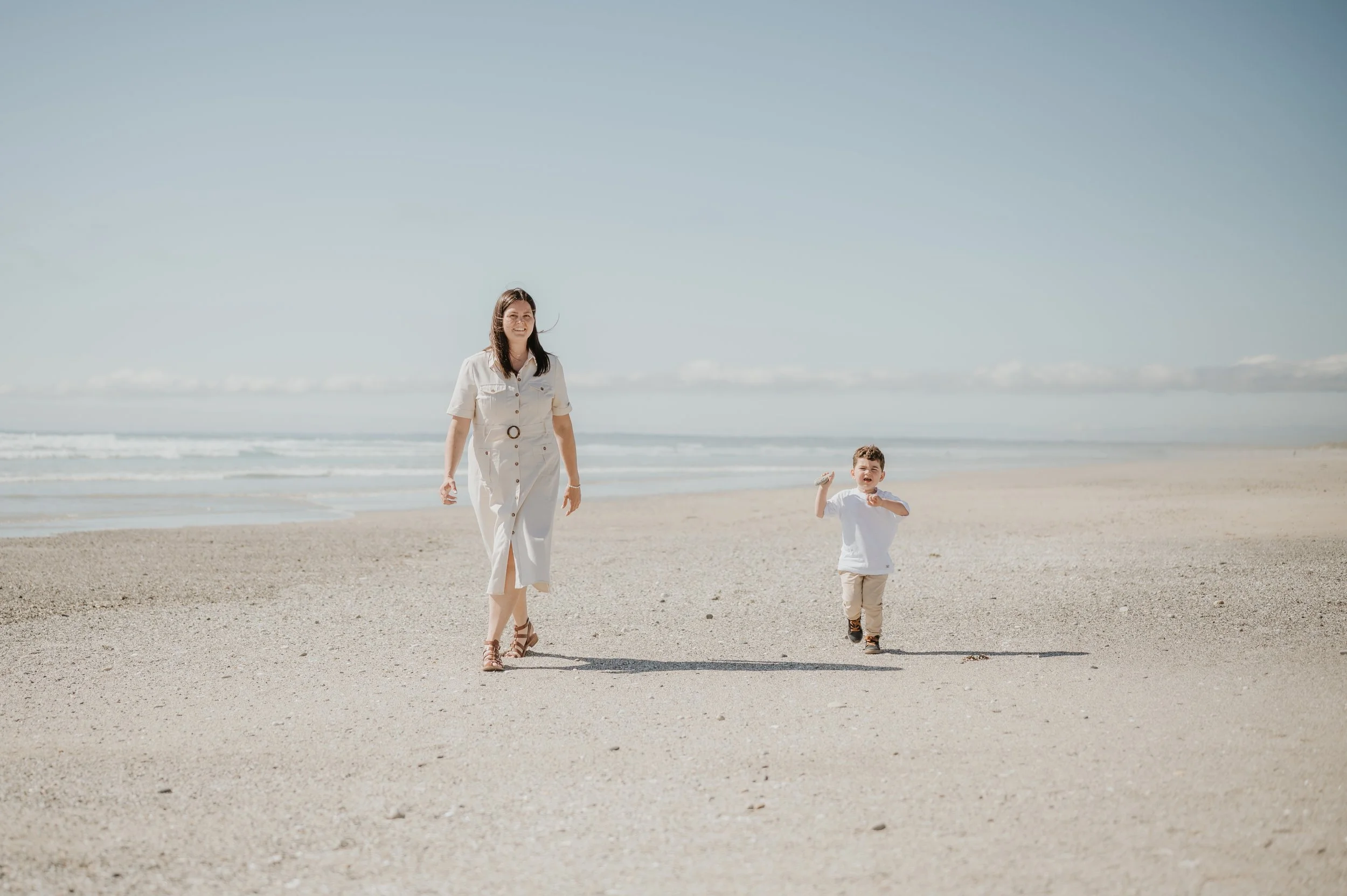 Une femme et un jeune garçon marchent sur la plage sous un ciel bleu avec peu de nuages.