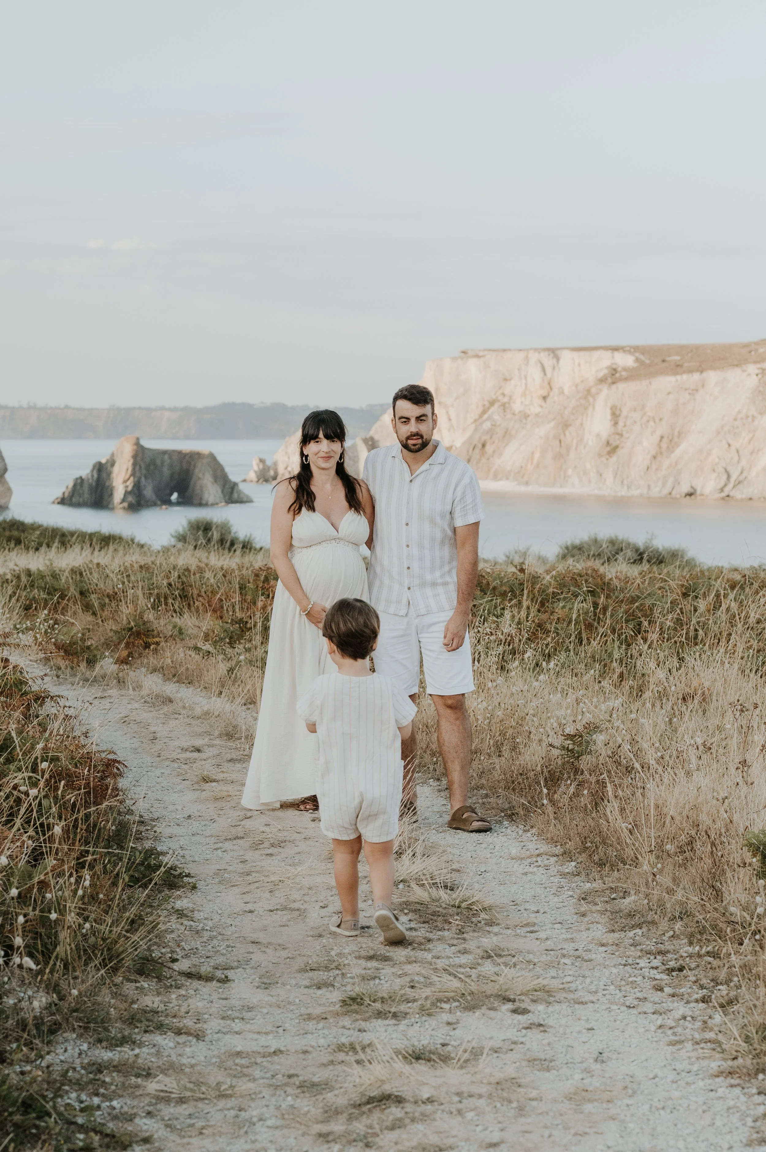 Une famille de 3 personnes, deux adultes et un enfant, se promènent sur un sentier de plage en direction de la mer avec des falaises en arrière-plan, dans un paysage côtier naturel au coucher du soleil.