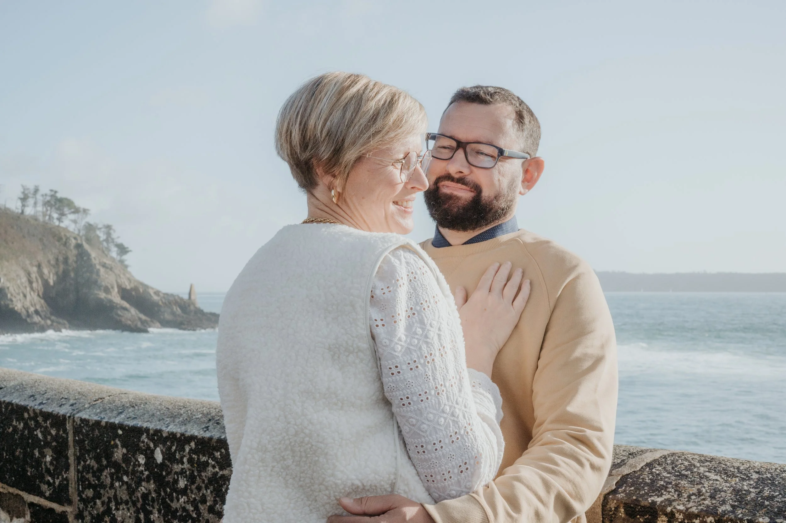 Un homme et une femme âgée qui se tiennent près de la mer, se regardant et souriant, avec un rocher en pierre devant eux et une côte rocheuse en arrière-plan, par une journée ensoleillée.