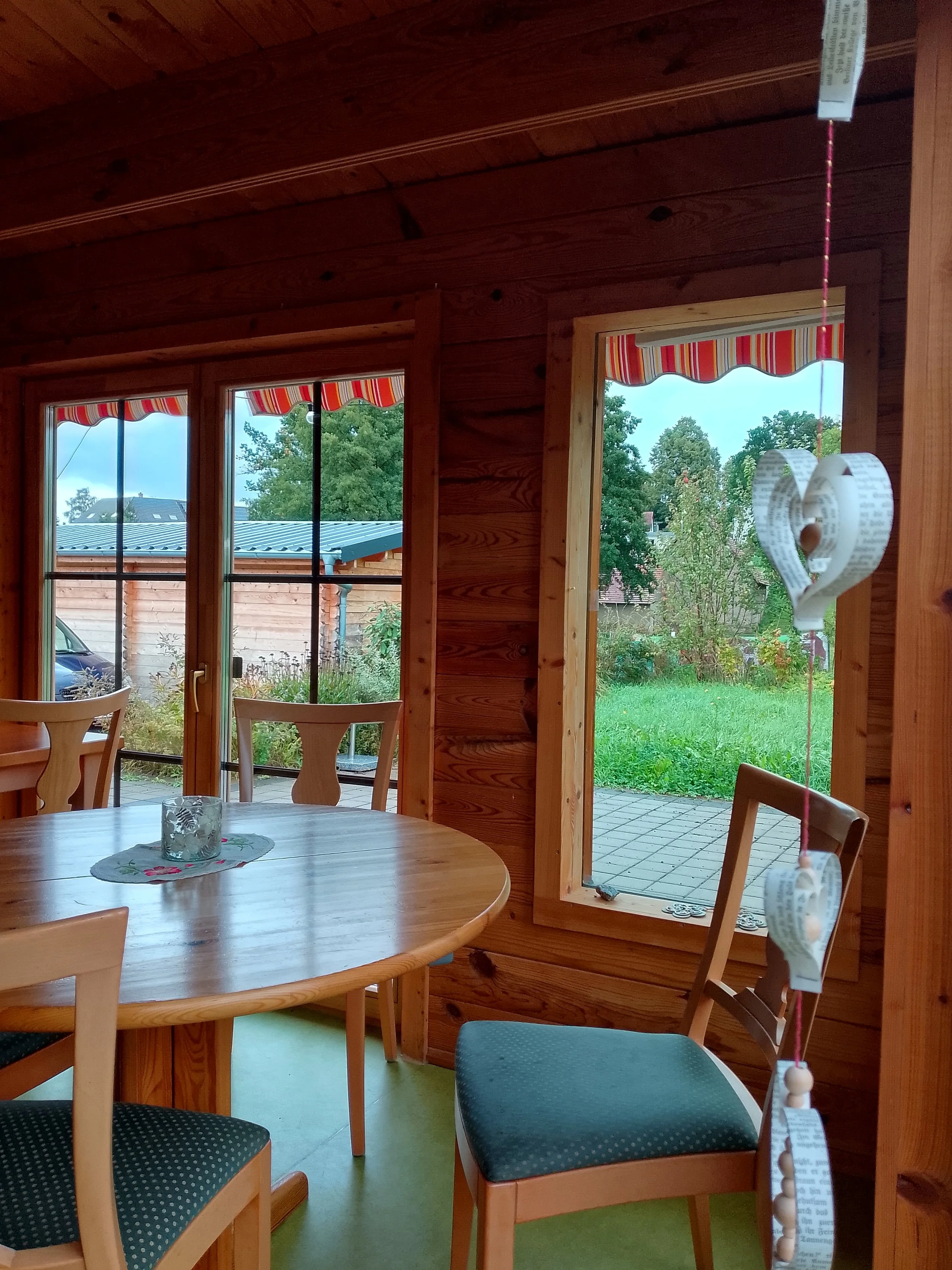 A cozy interior room with wooden walls and ceiling. There are three large windows with striped red, yellow, and white awnings. A round wooden dining table with a glass candle holder and a decorative cloth is in the foreground, surrounded by wooden chairs with upholstered seats. Outside, a garden with green grass, and trees, and a patio with a house and parked car are visible.