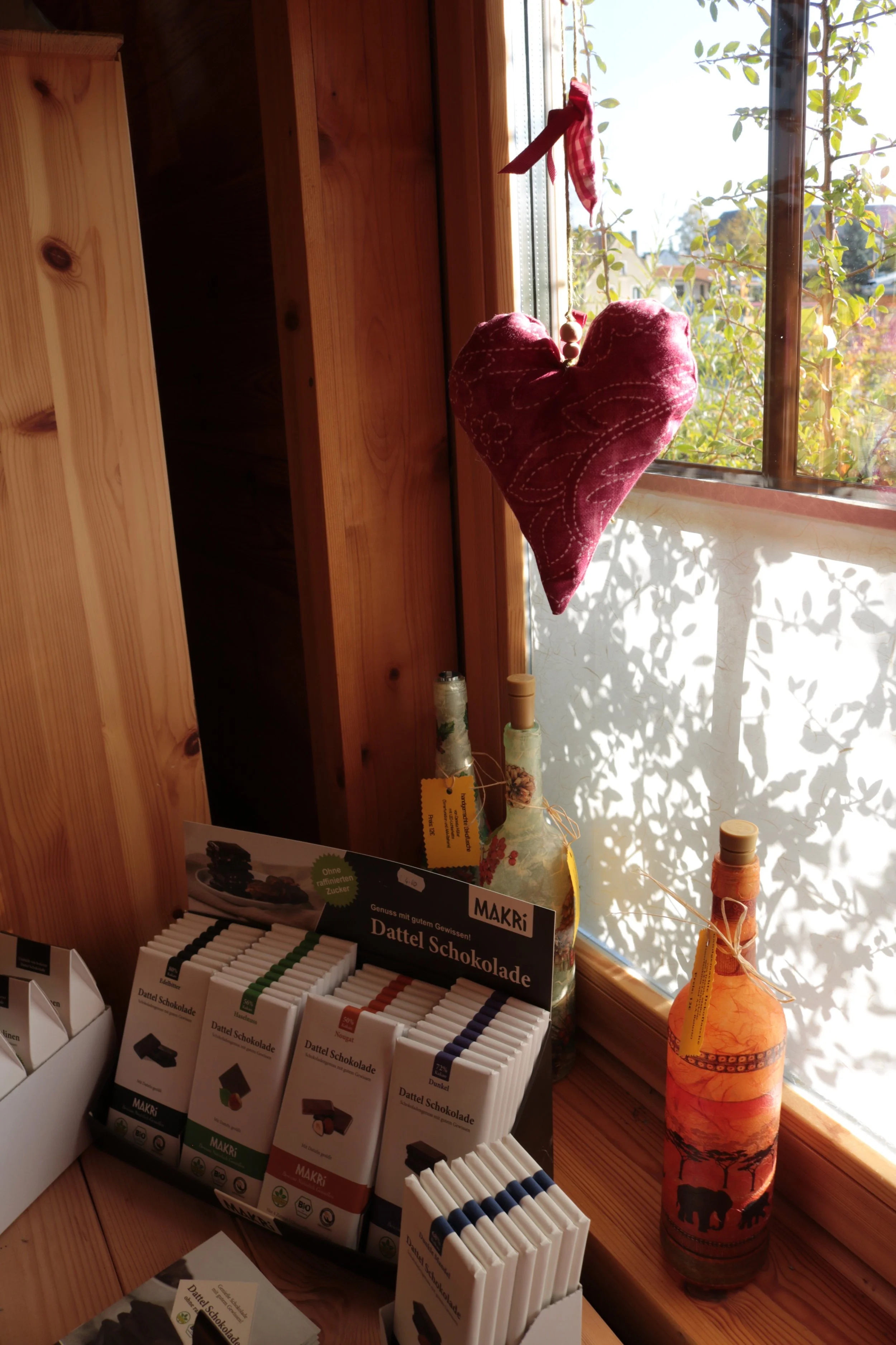 A red fabric heart ornament hanging by a string near a window with sunlight and green leaves outside. Several bottles and chocolate products are on a wooden surface below, with a display of Dattel Schokolade (date chocolate) bars in the foreground.