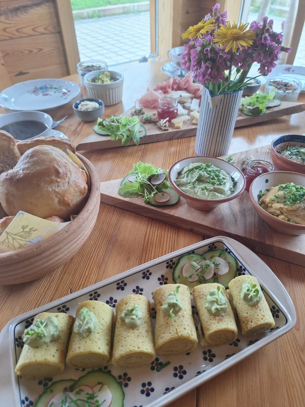 A table set with various breakfast foods including bread rolls, cucumber and radish slices, rolled omelets with green garnish, bowls with spreads or dips, a flower arrangement in a striped vase, and a charcuterie board with cheese, meats, and crackers.