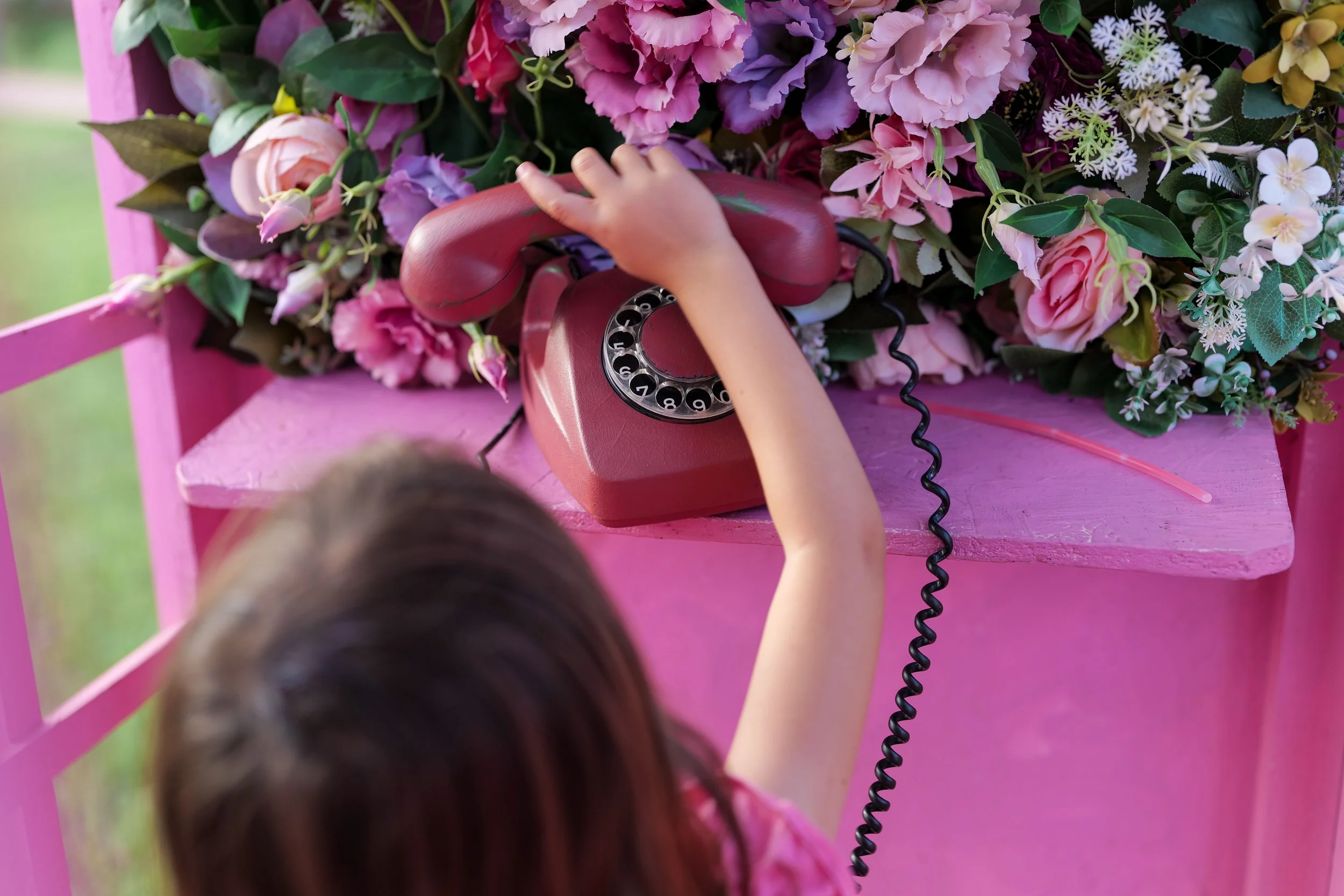 A child holding a pink rotary phone to a floral arrangement on a pink table.