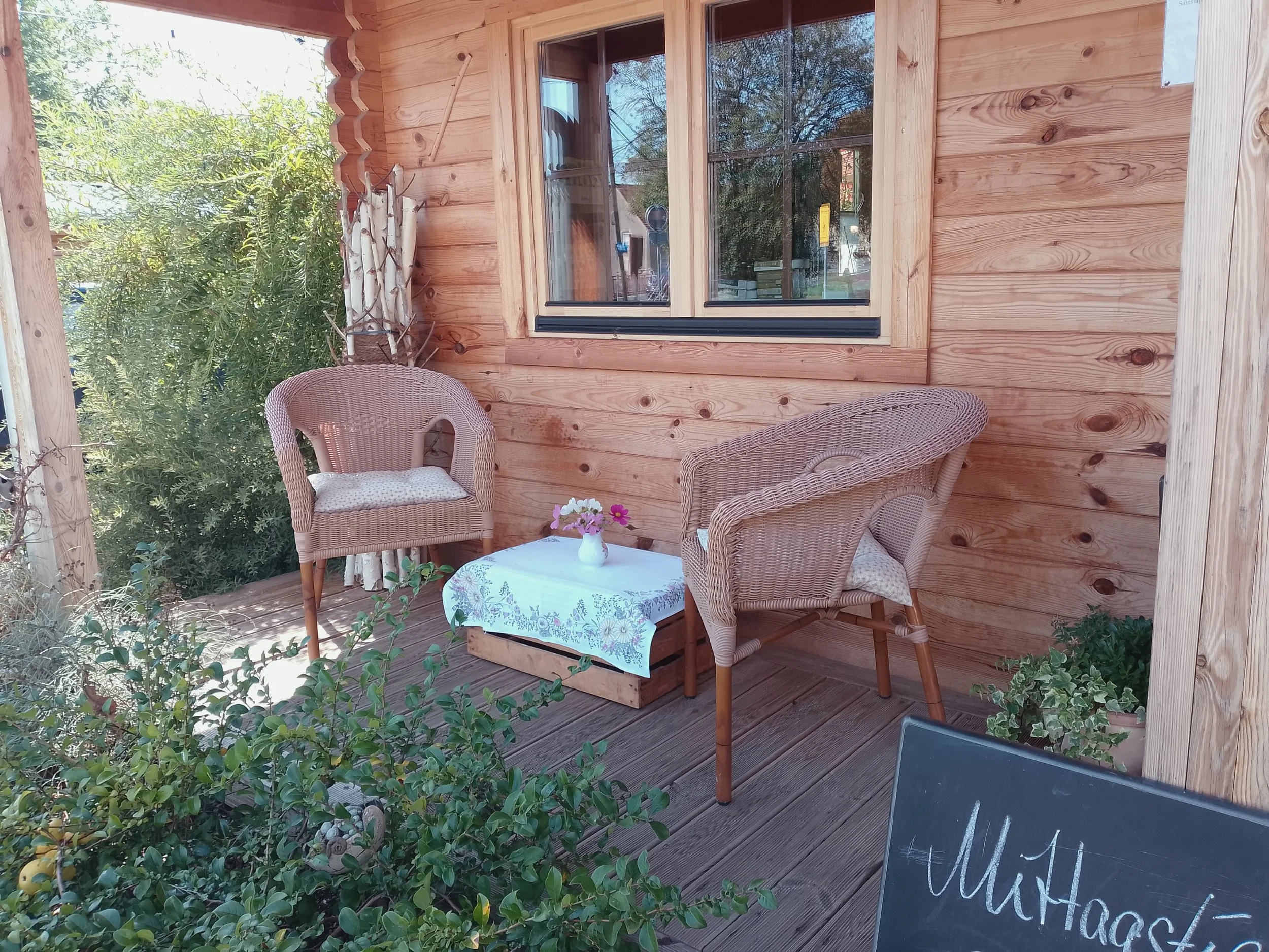 A cozy outdoor patio with wooden walls, wicker chairs with cushions, a table with a floral tablecloth and small flower vase, and a blackboard sign with handwritten text, surrounded by greenery.