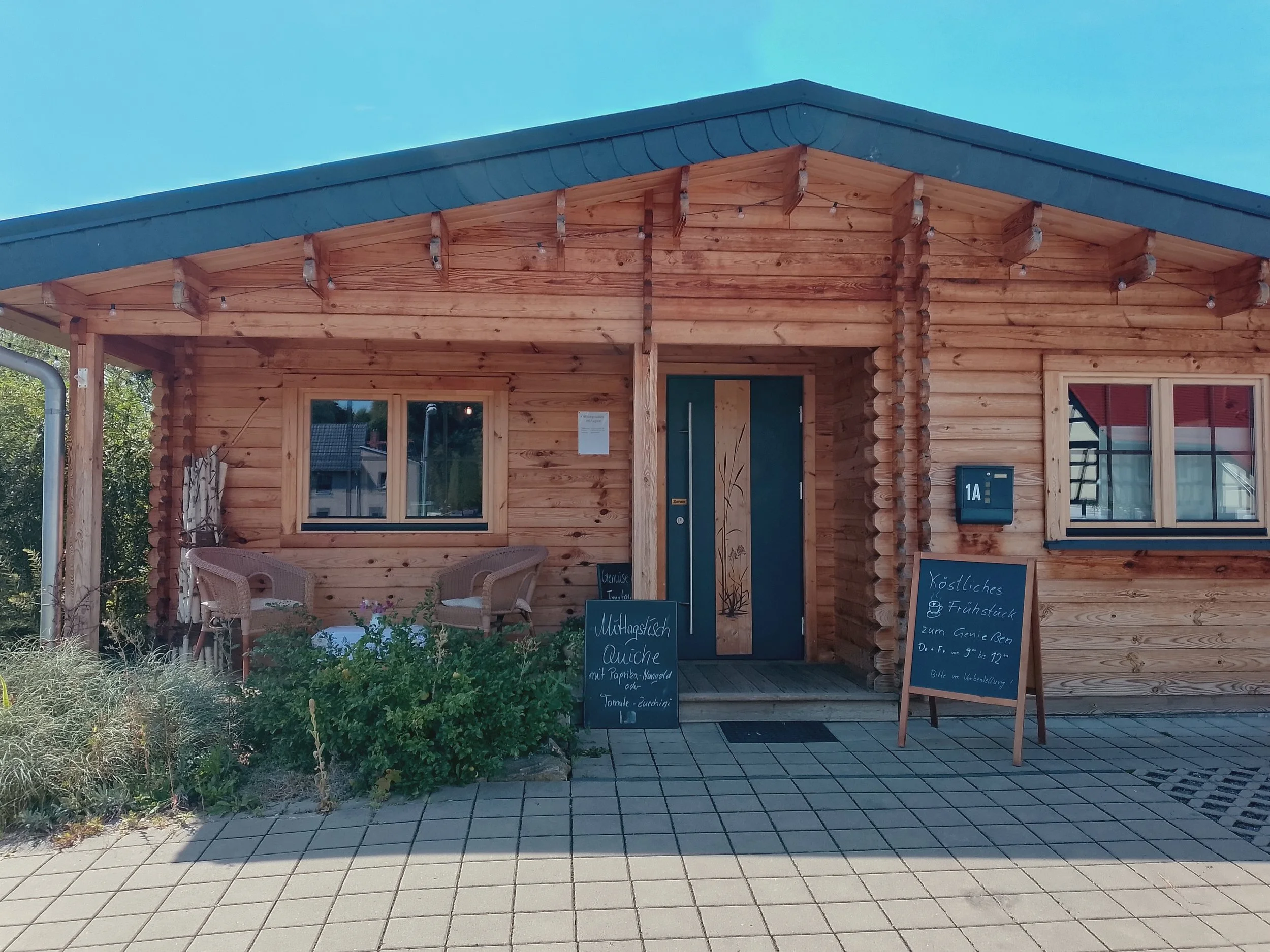 Front view of a wooden house with string lights hanging under the roof eaves, outdoor seating area with wicker chairs and plants, and menu boards placed in front of the entrance.