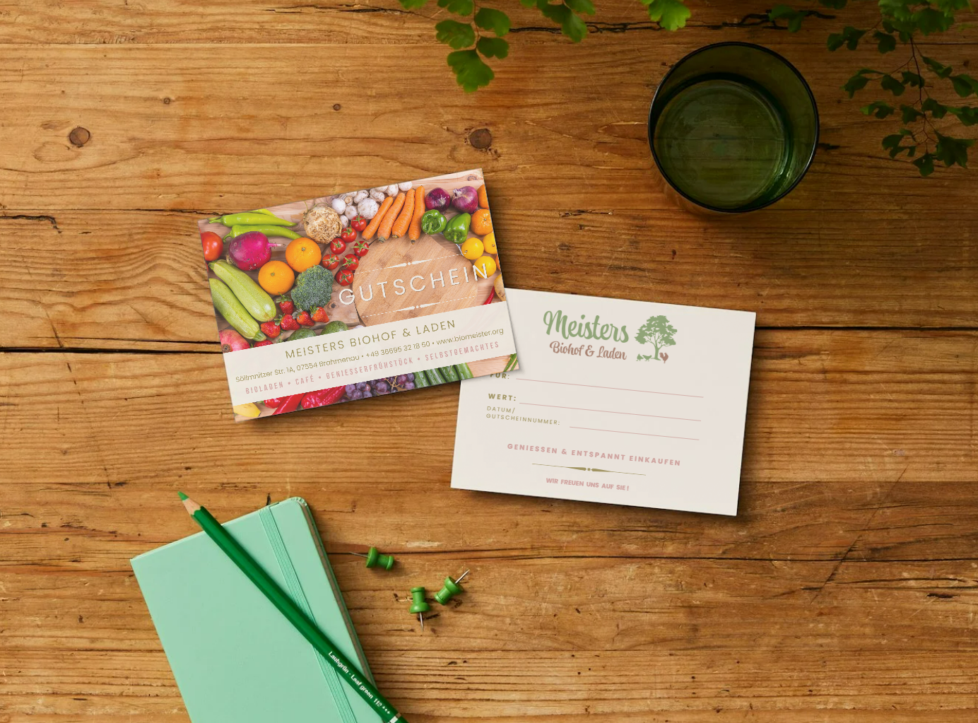 A wooden table with a green glass, two grocery gift cards, a light green notebook, a green pen, and three green push pins arranged on it.