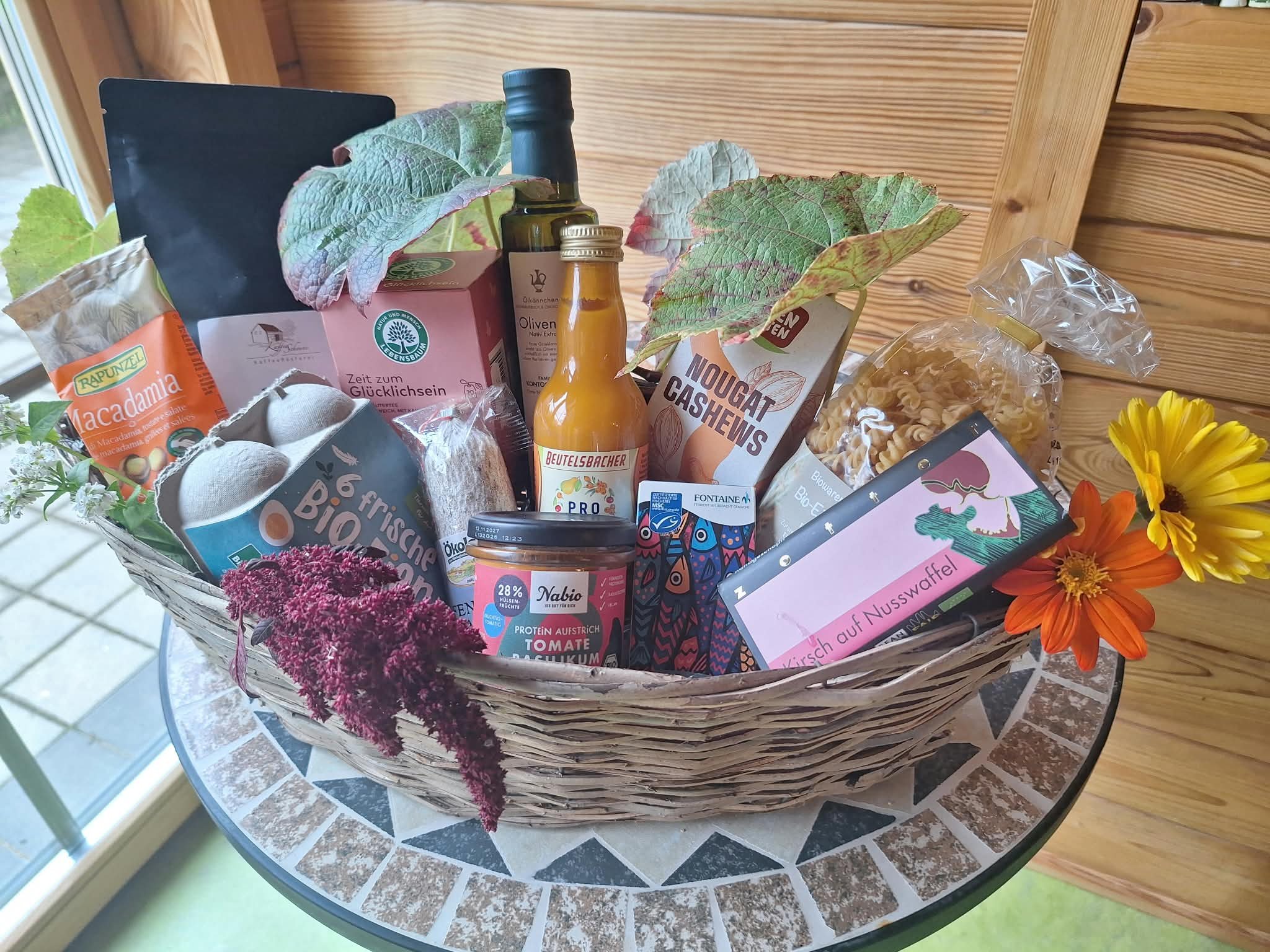 A wicker basket filled with various food items, drinks, and flowers, sitting on a glass table near a window with wooden walls in the background.