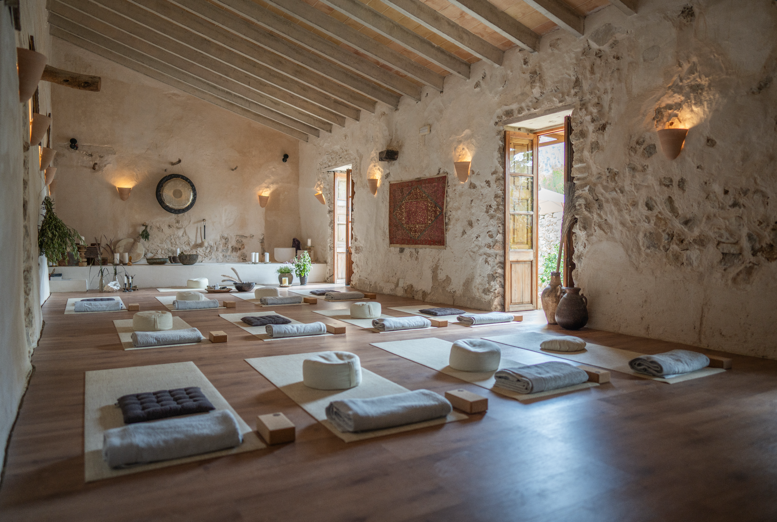 A serene yoga studio with stone walls, wooden ceiling beams, and large windows letting in natural light. Yoga mats, bolsters, and blocks are arranged on the wooden floor, creating a peaceful setting for a class or meditation.