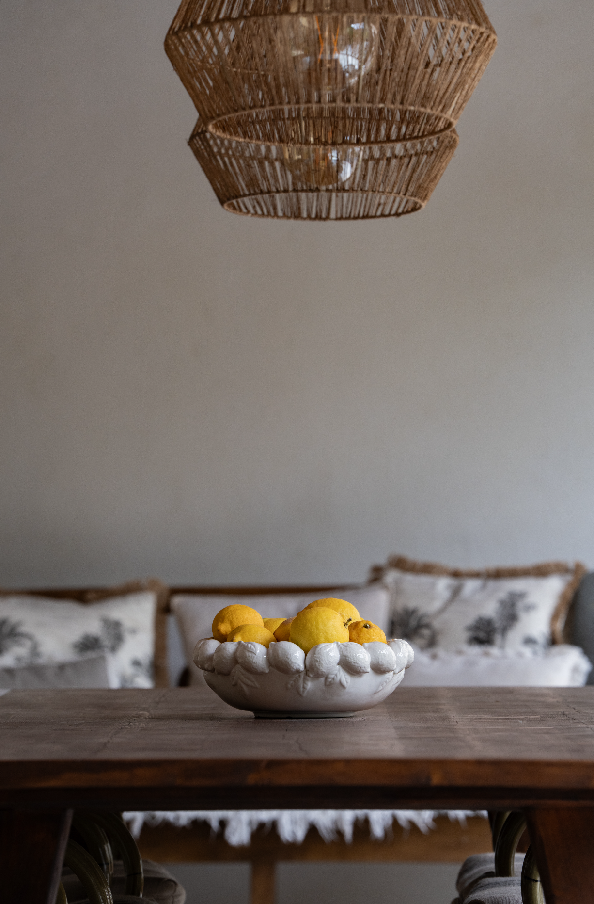 A white ceramic bowl filled with yellow lemons sits on a wooden dining table. In the background, there are decorative pillows with black and white floral patterns on a seating area, and a wicker lampshade hangs from the ceiling.