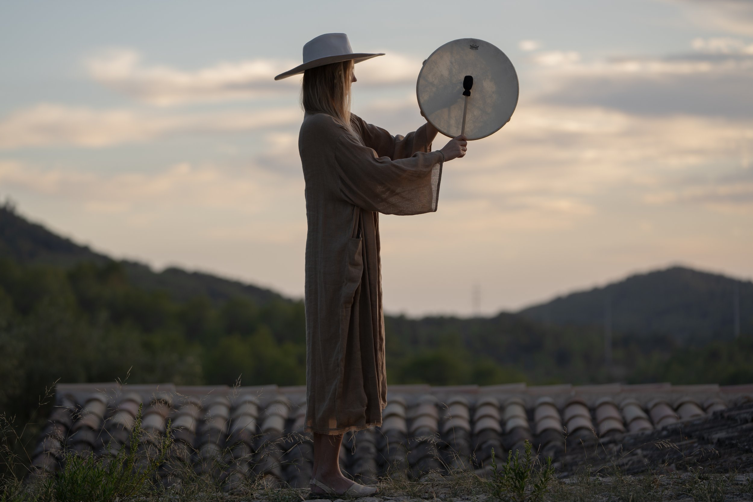 A woman with long hair wearing a wide-brimmed hat and a loose dress standing on a rooftop at sunset, holding a circular reflector.