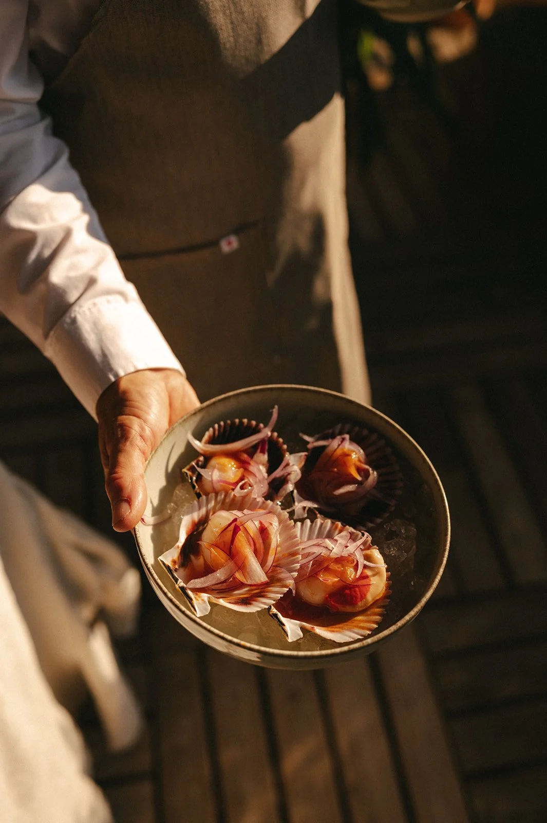 A person holding a bowl of fresh seafood, including scallops and shells, on a dark patio.