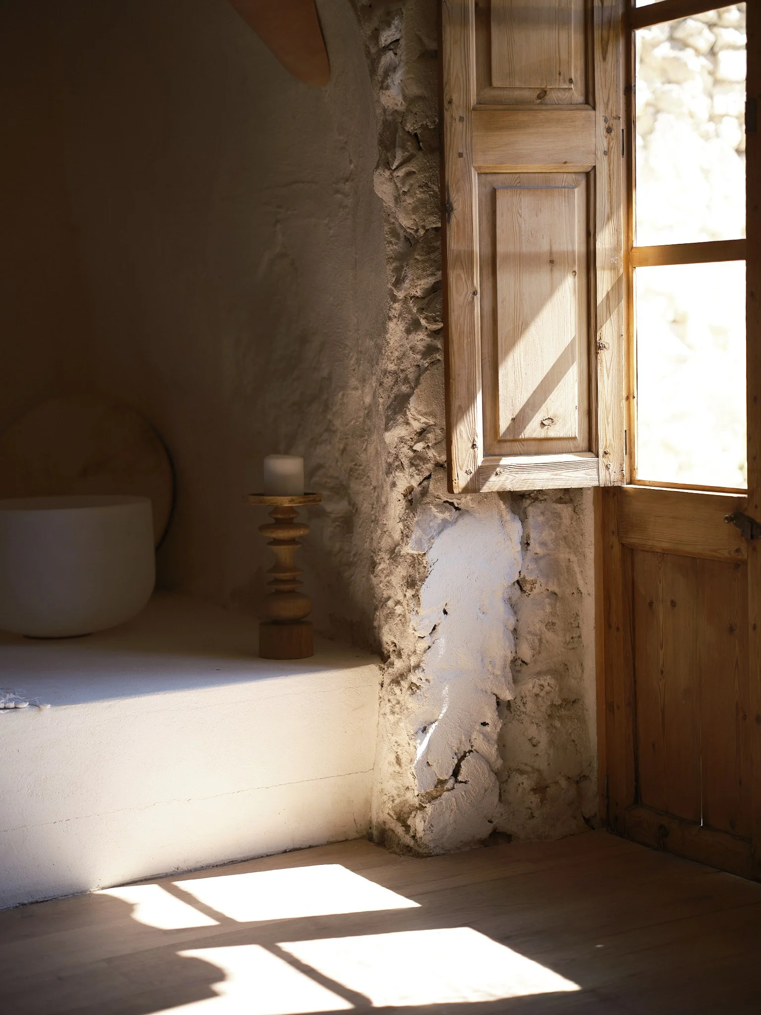 Open wooden window with sunlight casting shadows on a rustic stone wall inside a room, with a small white candle on a wooden stand and a neutral-colored lamp on a white wall.