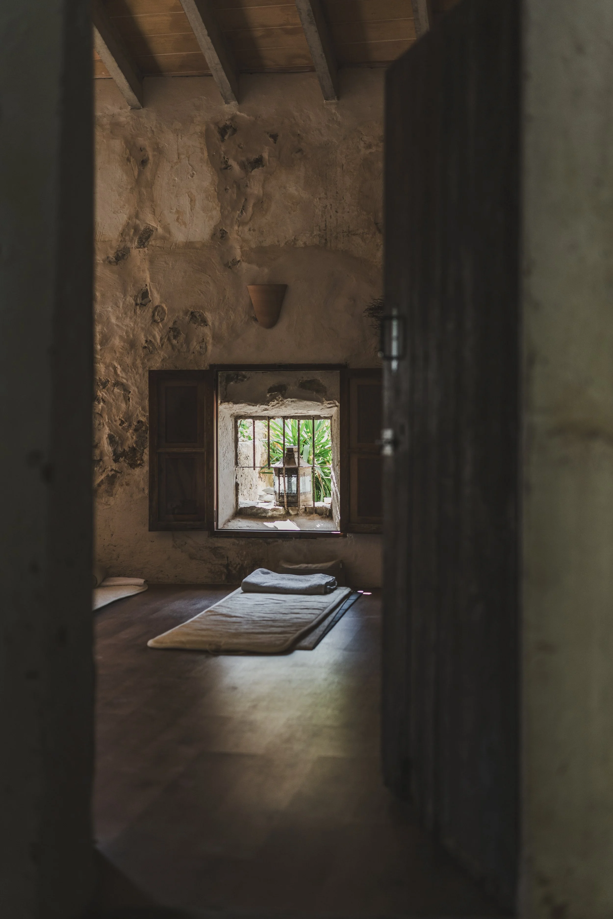 A cozy rustic room with exposed stone walls, a wooden ceiling, a small window with shutters, a lantern on the window sill, and a rug with folded towels on the wooden floor.