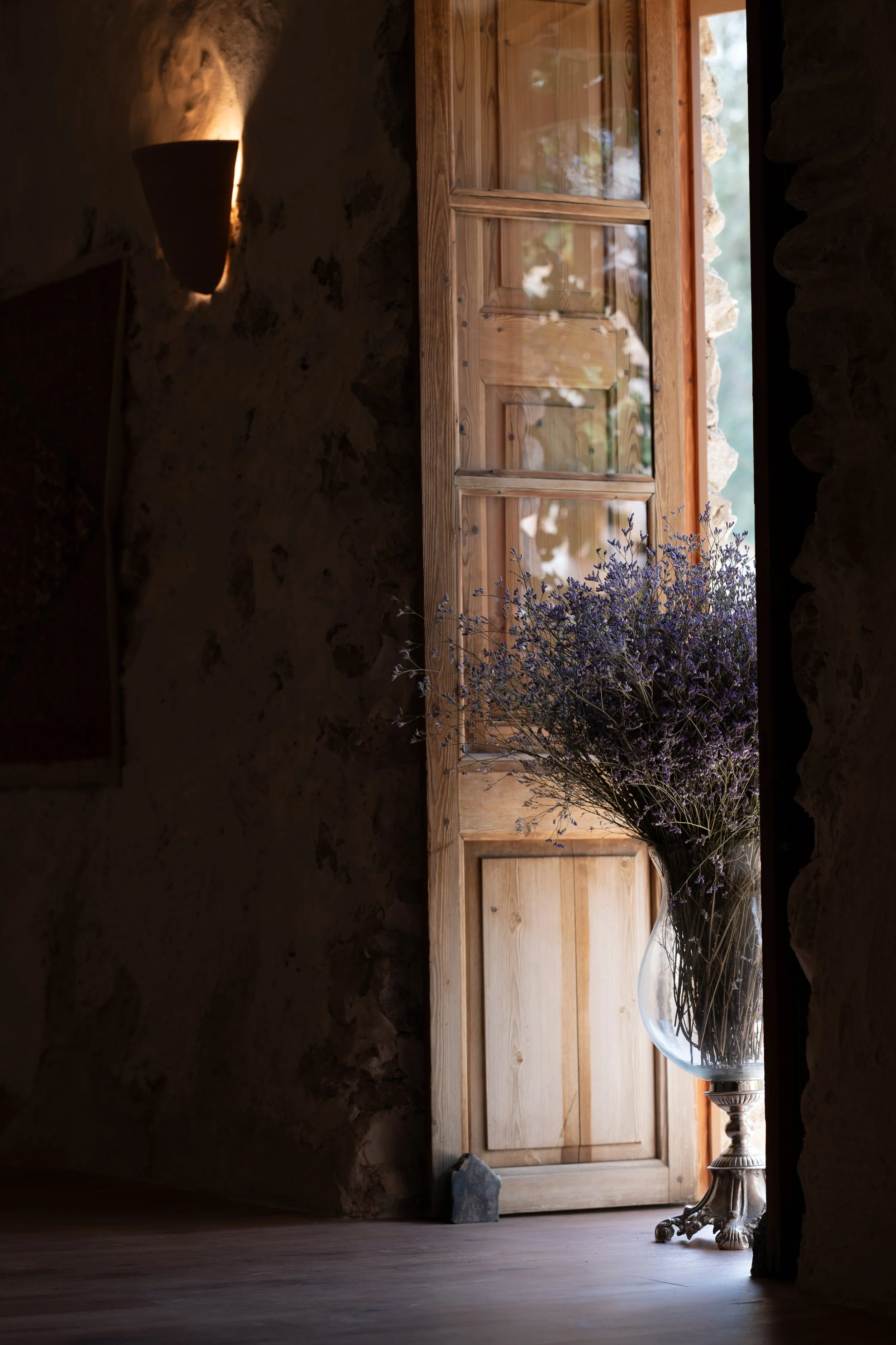 A wooden door with a glass window, set into a stone wall, with a vase of purple flowers and a small stone on the wooden floor near the door, lit by natural light.