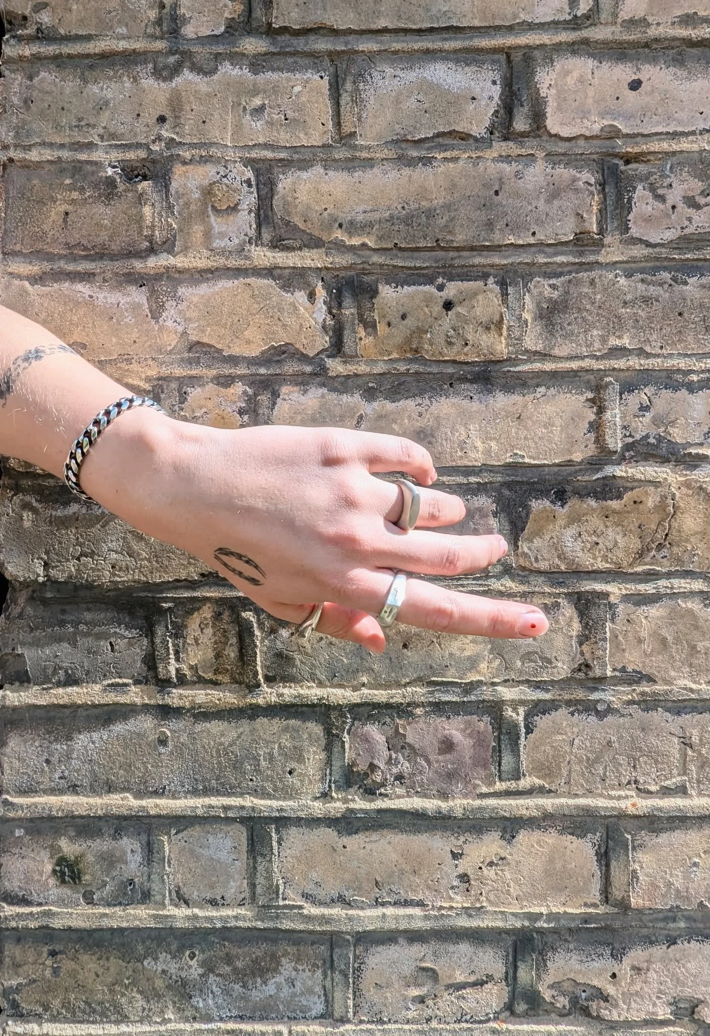 A person's hand with rings and bracelets extended horizontally against a weathered brick wall.