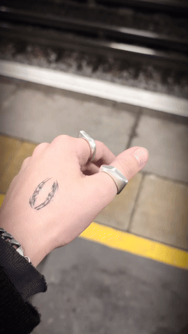 A hand with rings and a tattoo, with train tracks and platform in the background.