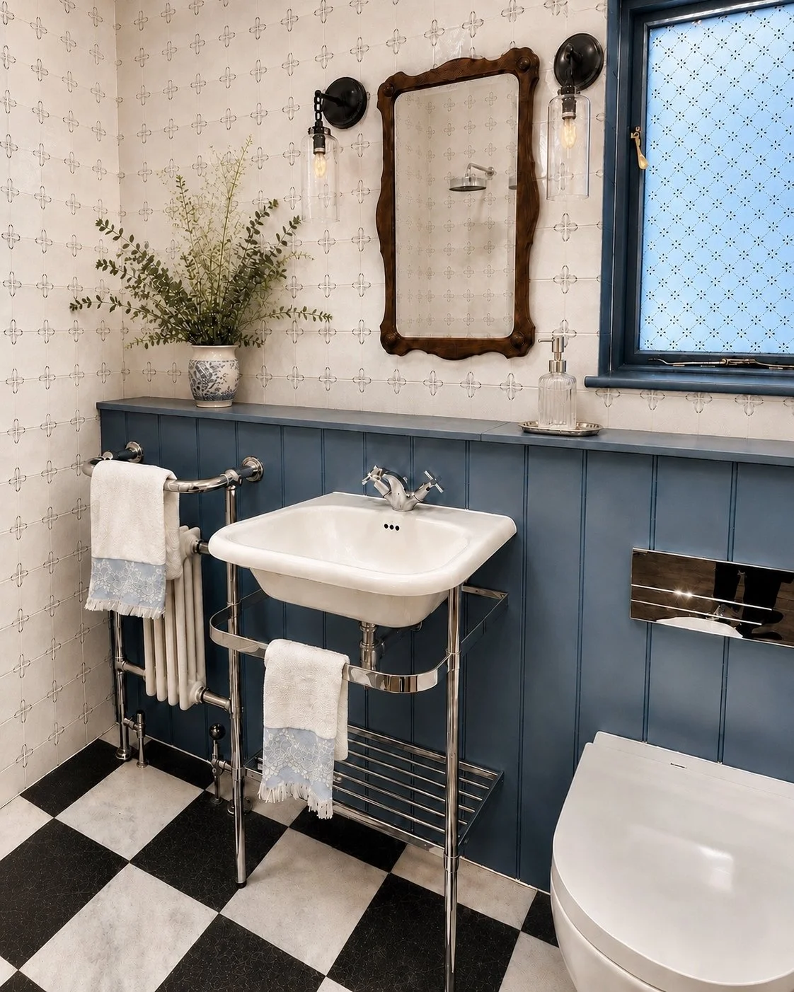 A vintage-style bathroom with a blue wainscot panel behind a white sink, black and white checkered floor, a black wall-mounted mirror, and a window with frosted glass. There is a vase with greenery, a soap dispenser, and wall-mounted light fixtures.