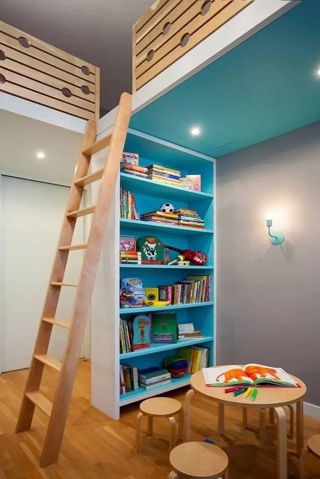 Kids' room with a bookcase, angled wooden ladder, and a lofted play area. The bookcase is filled with children's books and toys. There is a round table with coloring supplies and small stools on a wooden floor.