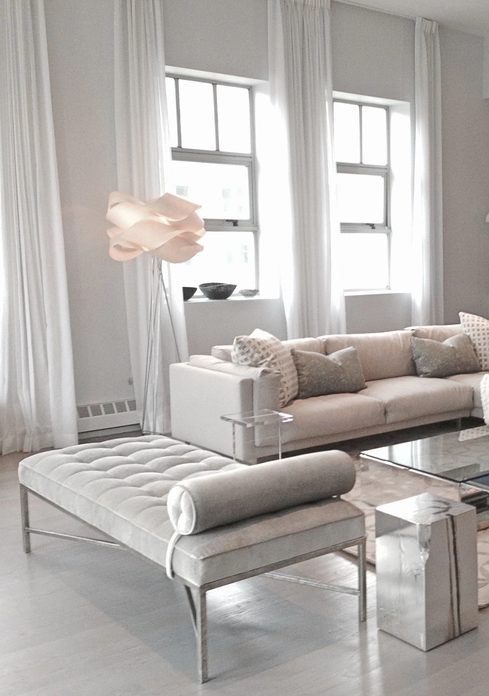 A modern living room with light-colored walls, large windows with white curtains, a beige sofa with pillows, a white upholstered bench, and a glass coffee table. There is a decorative floor lamp with a sculptural shade.