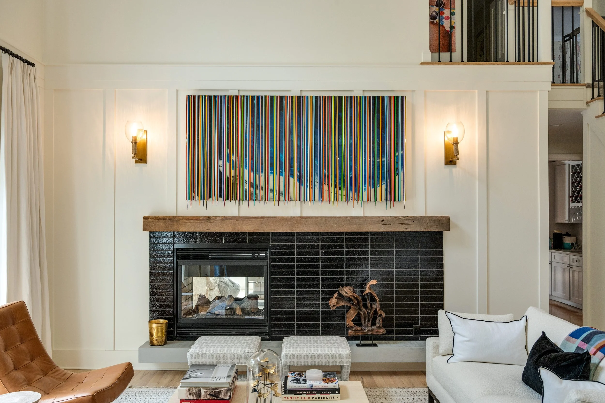 Living room with a black brick fireplace, colorful striped art above it, and modern furniture including a white sofa with pillows, a brown leather chair, and a glass coffee table with books and decorations.