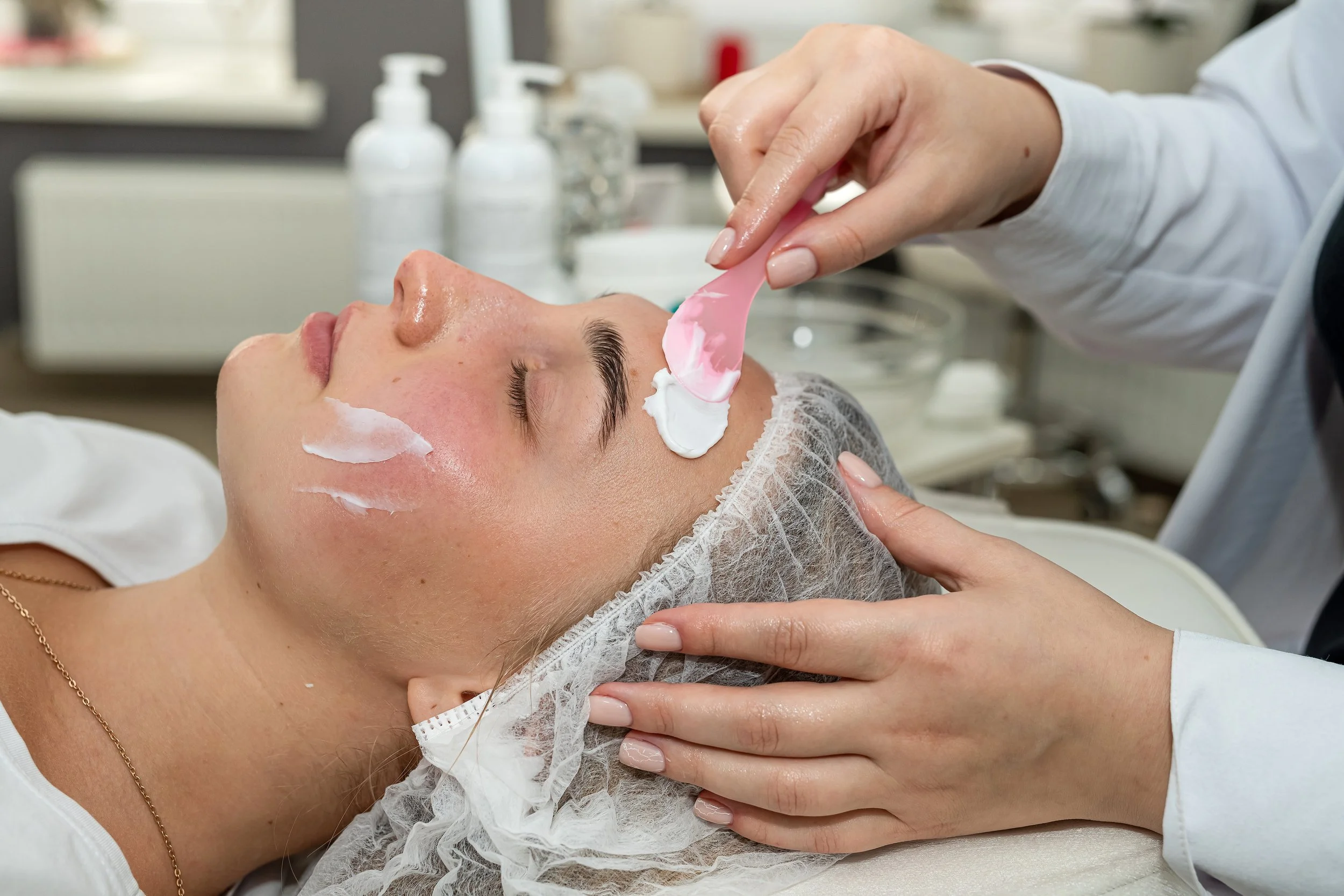 A woman receiving a facial treatment in a spa or skincare clinic, lying down with her eyes closed while a professional applies a skincare product with a pink spatula.