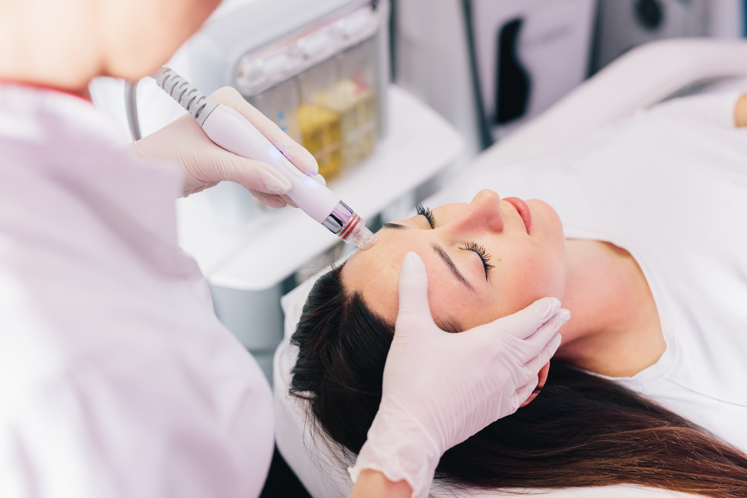 A woman lying down while a professional performs a facial treatment using a handheld device.