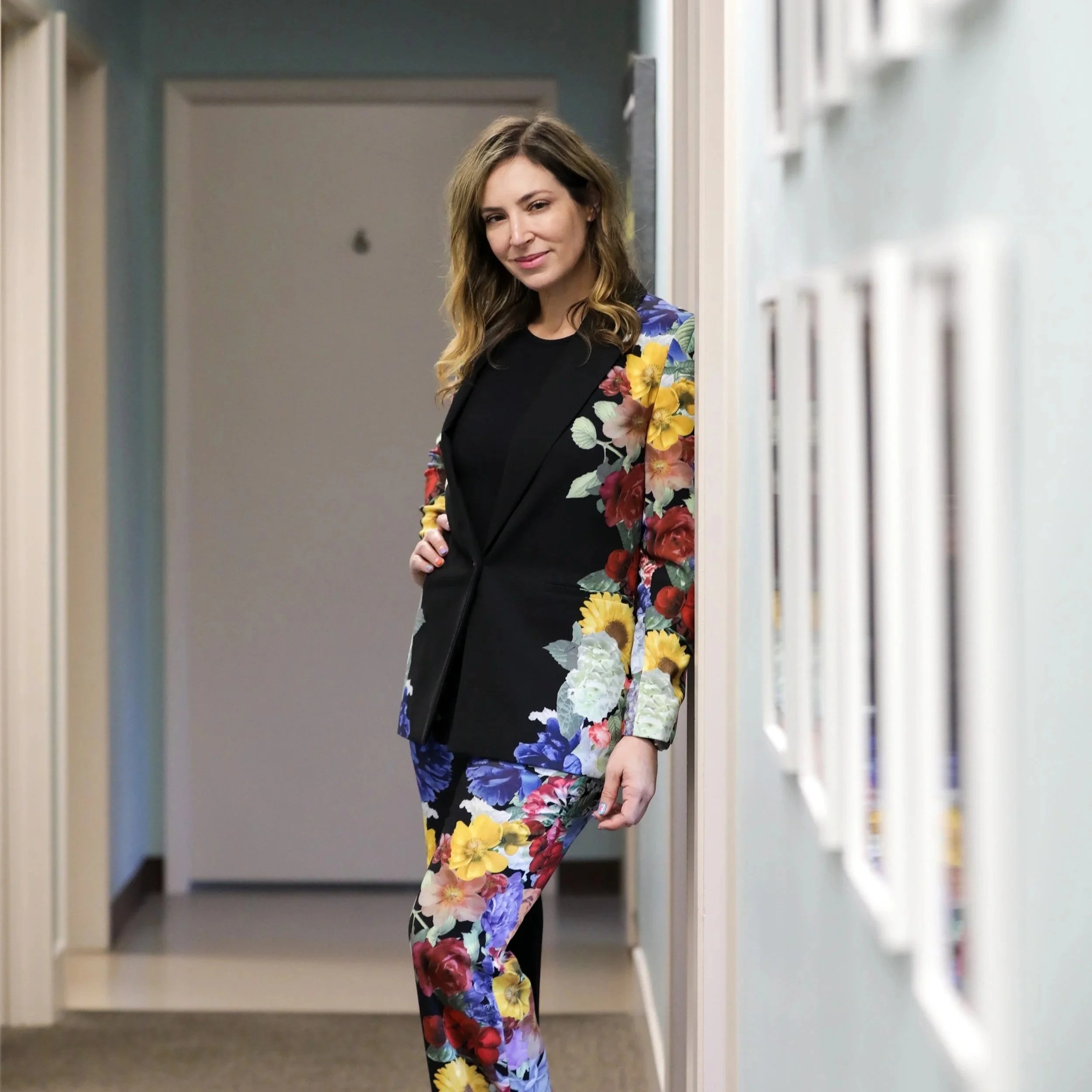 A woman with long, wavy brown hair wearing a black top and a vibrant floral blazer and matching skirt, standing in a hallway lined with bookshelves.