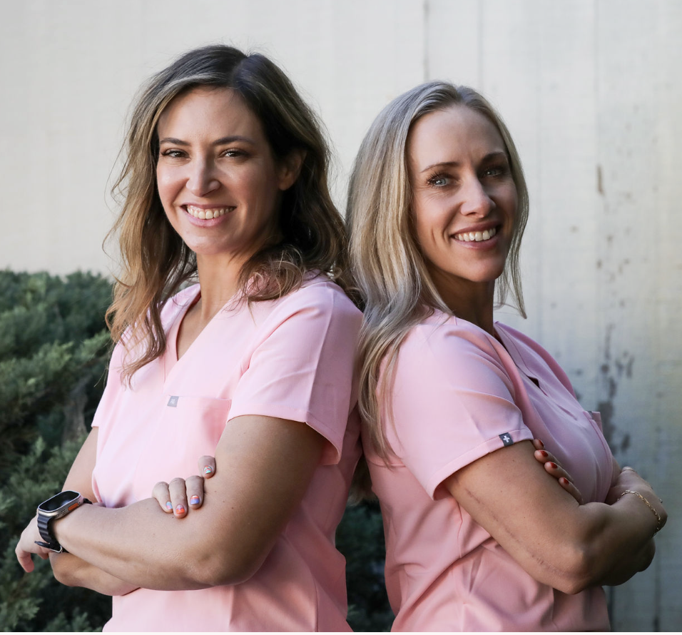 Two women wearing pink scrubs standing outdoors with arms crossed, smiling and posing back-to-back.