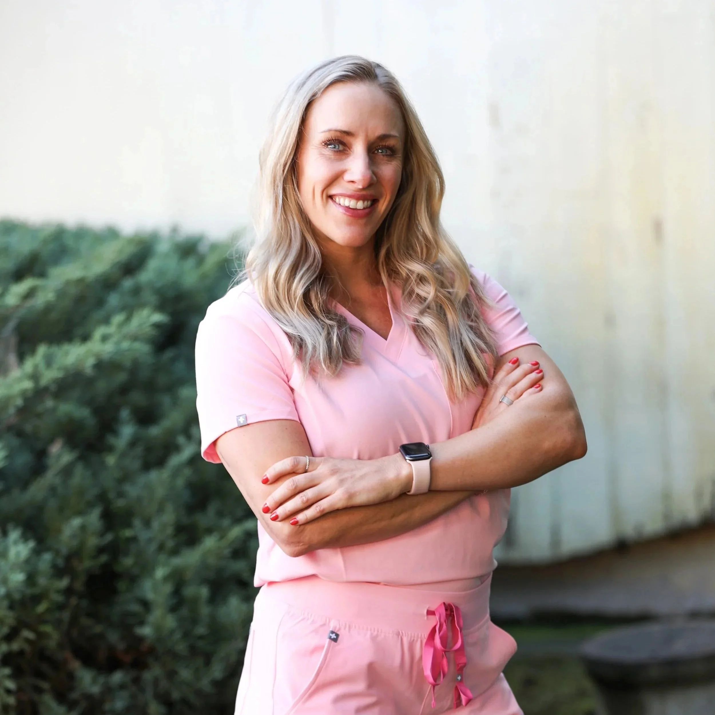 A woman with long blonde hair smiling, wearing a light pink outfit with short sleeves, standing outdoors in front of green foliage and a white wall.