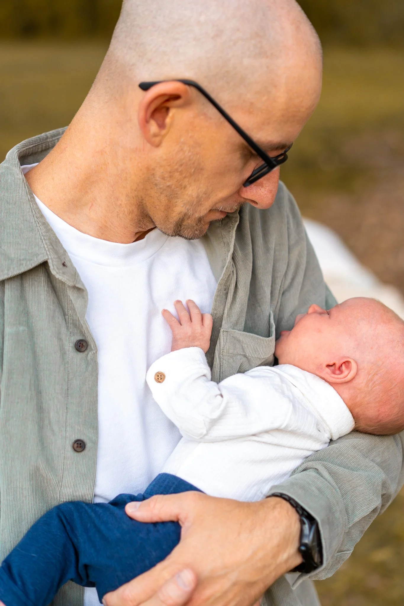 A man with glasses and a beard holds a baby outside. The man is looking down at the baby, who is reaching up to touch his face.