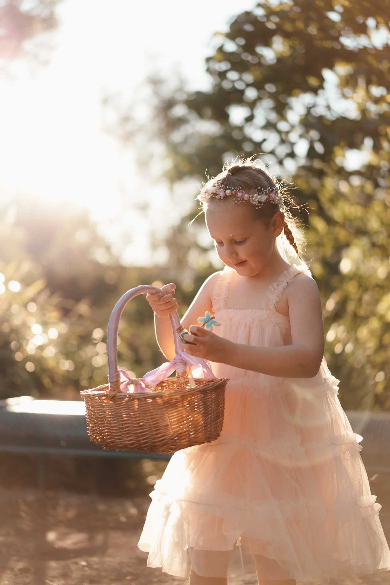 A young girl in a peach dress and floral crown holding a wicker basket outdoors during sunset with trees and sunlight in the background.