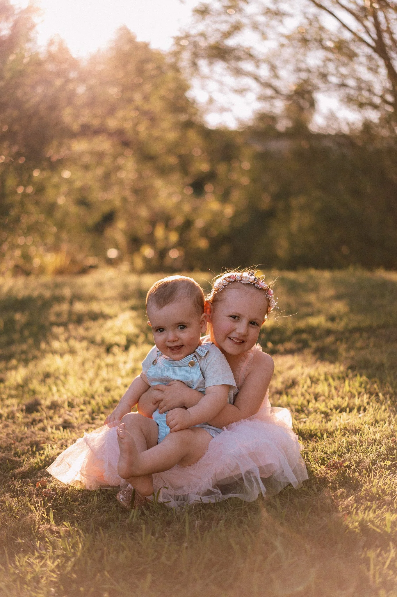 Two children, a girl and a boy, sitting on grass outdoors at sunset. The girl wears a pink dress and floral headband, smiling at the camera, while the boy wears a light blue shirt with suspenders, also smiling. The background includes blurred trees a
