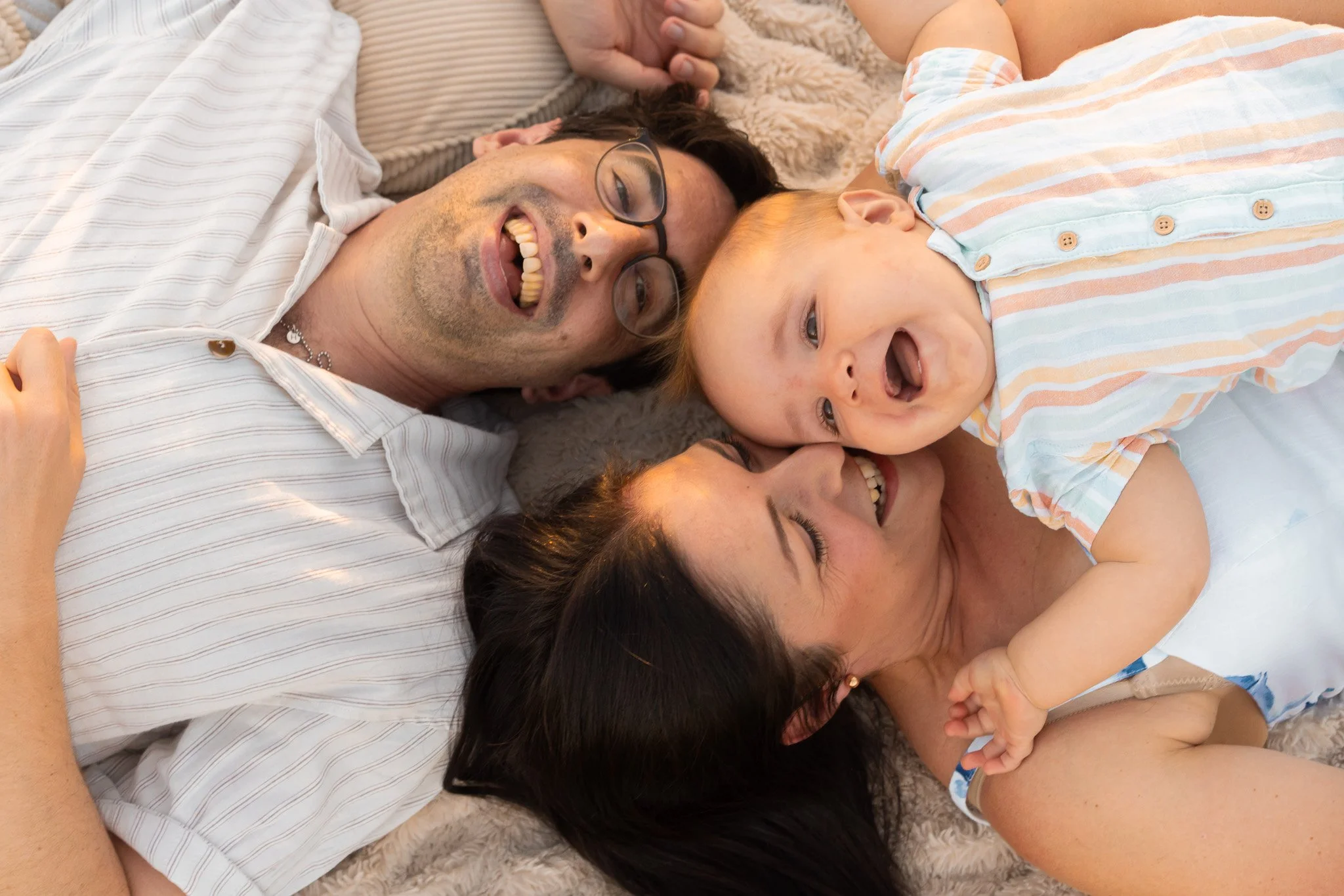 A family lying on a soft blanket, smiling and playing together. The man is wearing glasses and a striped shirt, the woman has dark hair, and the child has red hair and is wearing a striped shirt.