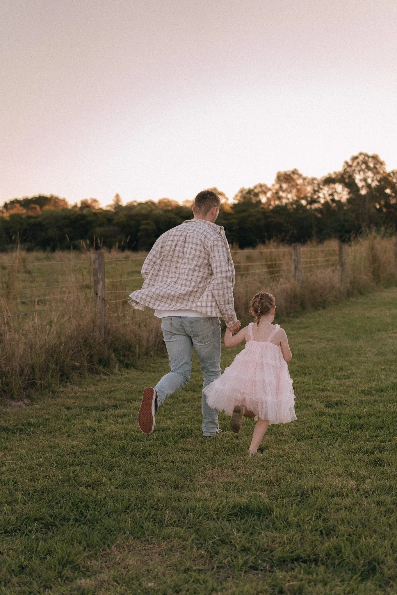 A man and a young girl holding hands and running across a grassy field near a fence during sunset.