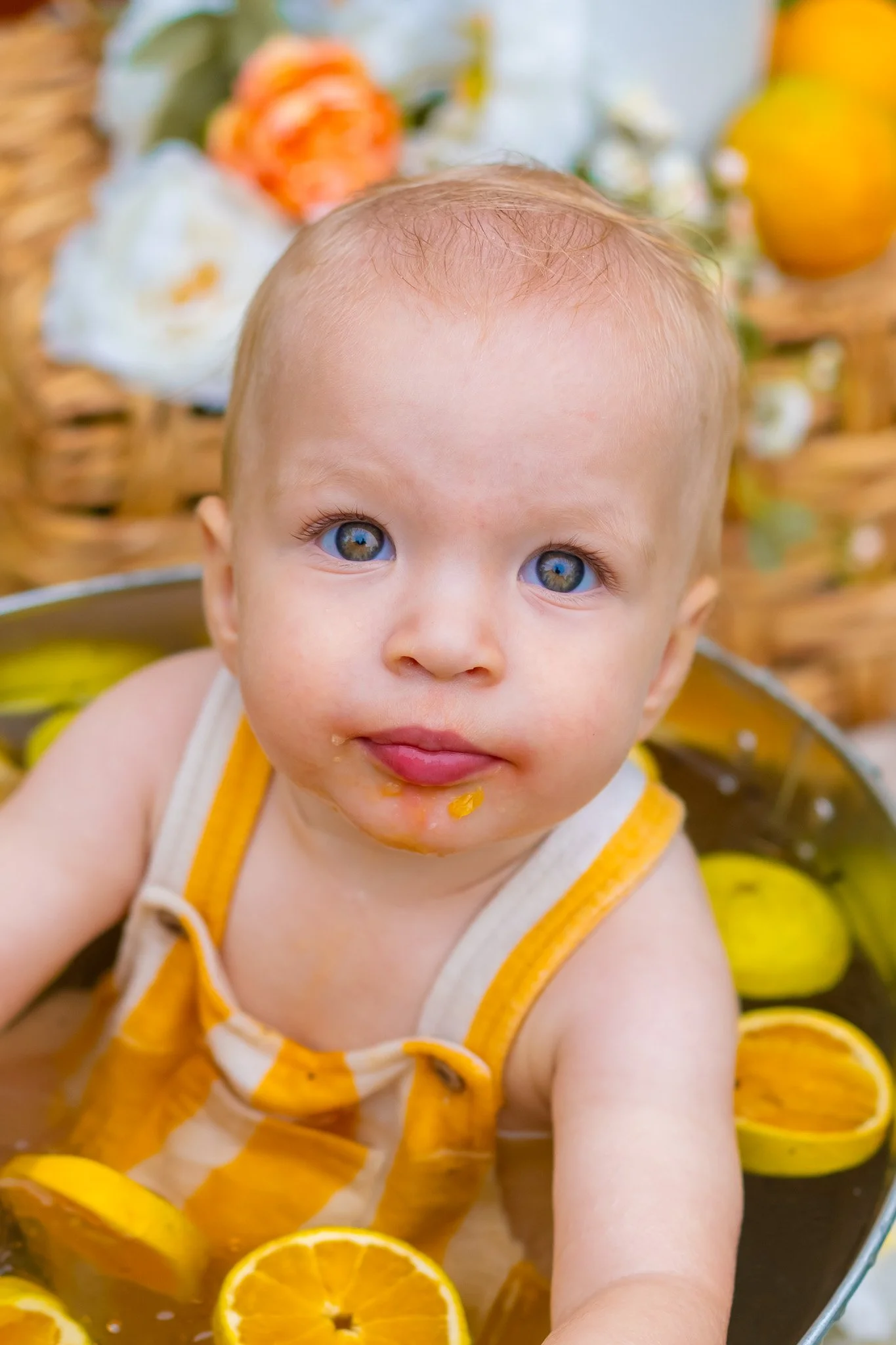 Close-up of a baby with blue eyes, wet hair, wearing a yellow and white striped sleeveless outfit, sitting among sliced orange oranges with a wicker basket and flowers in the background