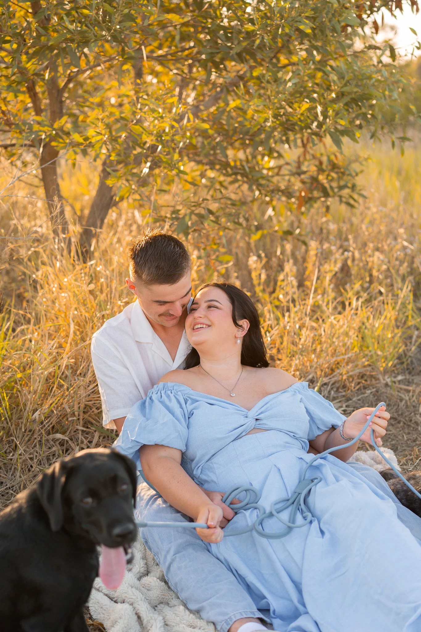 A couple sitting on the grass in a field with tall dry grass and trees during sunset. The woman is lying down in a light blue dress, holding a phone, and the man is sitting behind her, smiling, wearing a white shirt. A black dog is in the foreground,