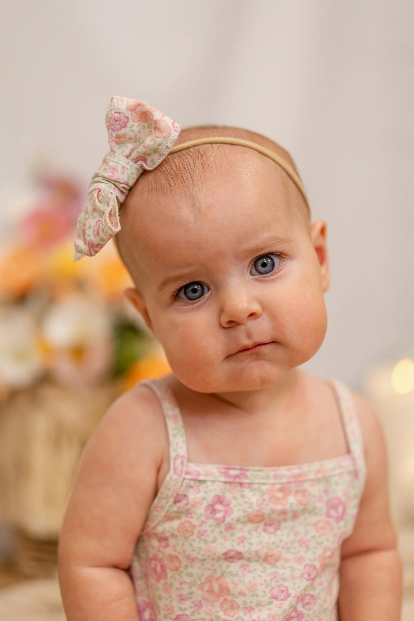 Close-up of a young girl with blue eyes and light skin wearing a floral-patterned dress and a headband with a bow, standing in front of a blurred background with soft-colored objects.