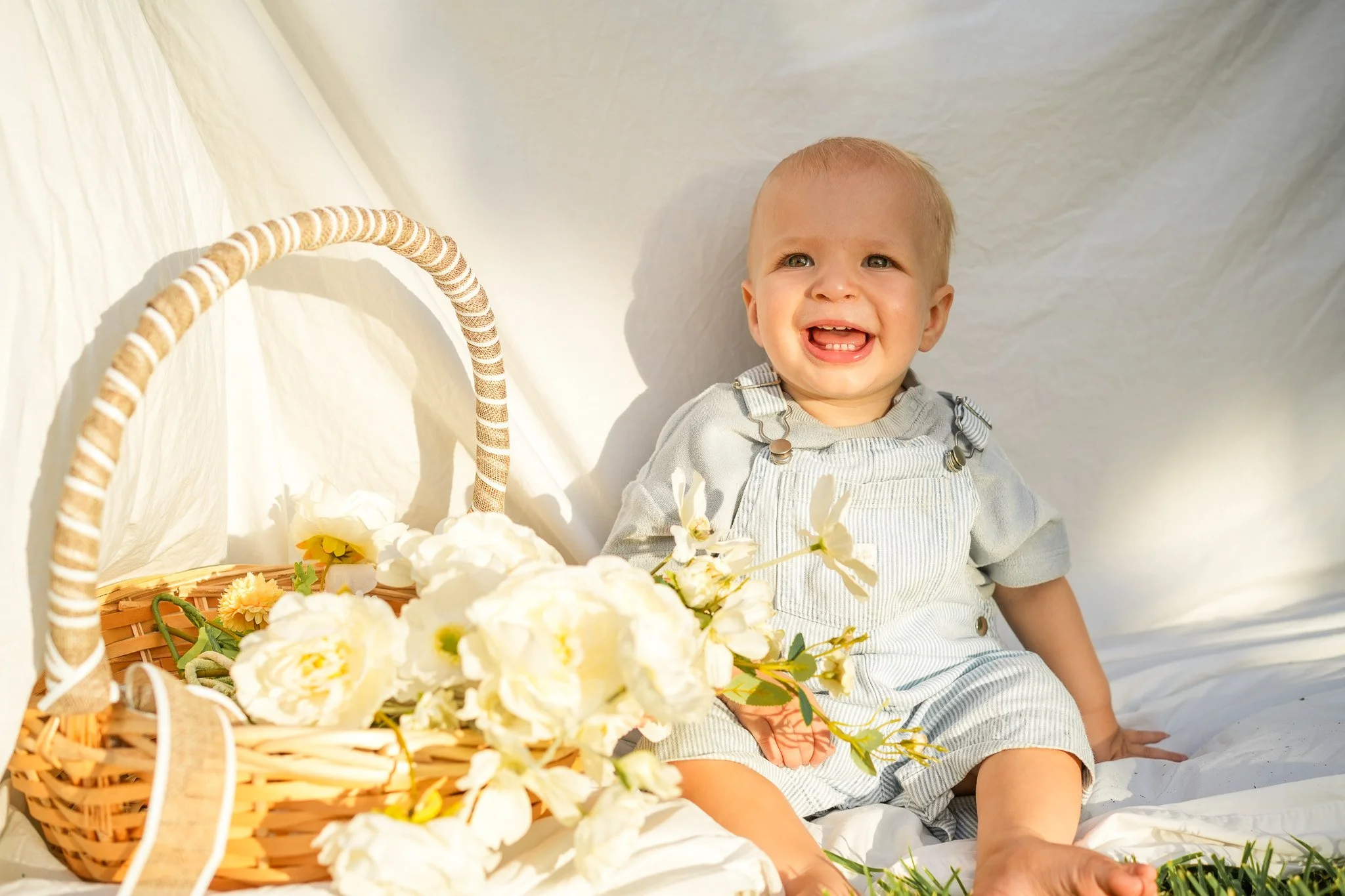 A smiling young child with blond hair sitting next to a basket of white and yellow flowers, on a white fabric background.