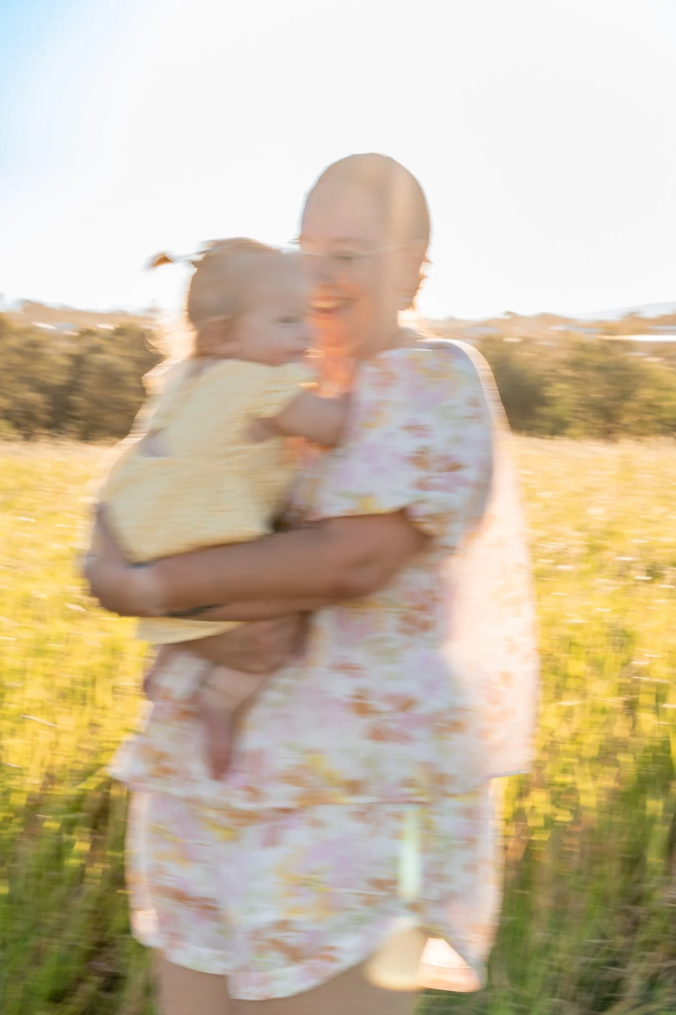 A person holding a young girl outdoors in a field with trees in the background on a sunny day.
