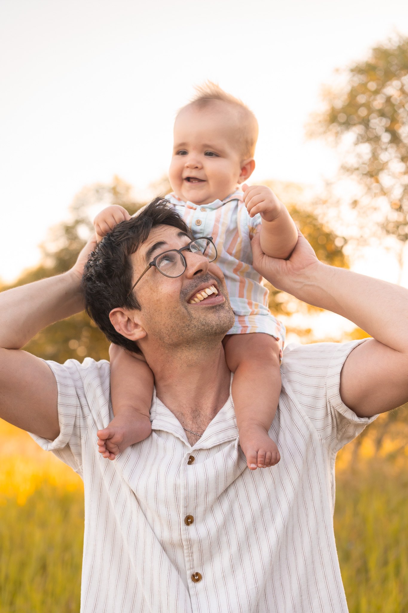 A man with glasses and a striped shirt is smiling up as a young child sits on his shoulders, outdoors in a field with trees and warm sunlight in the background.