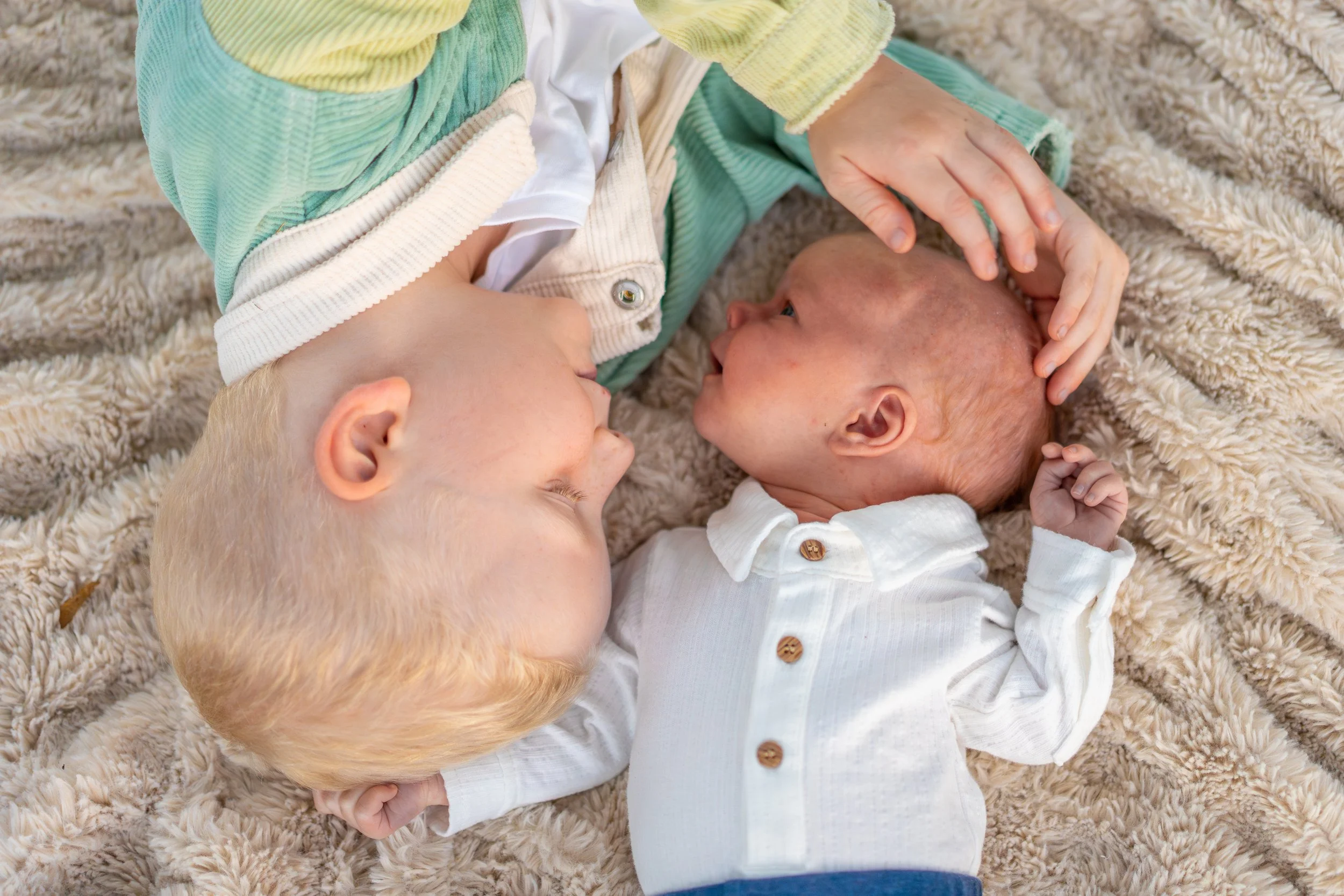 Two young children, one with blond hair and one with bald head, lying on a fluffy beige rug, facing each other, touching foreheads and smiling.