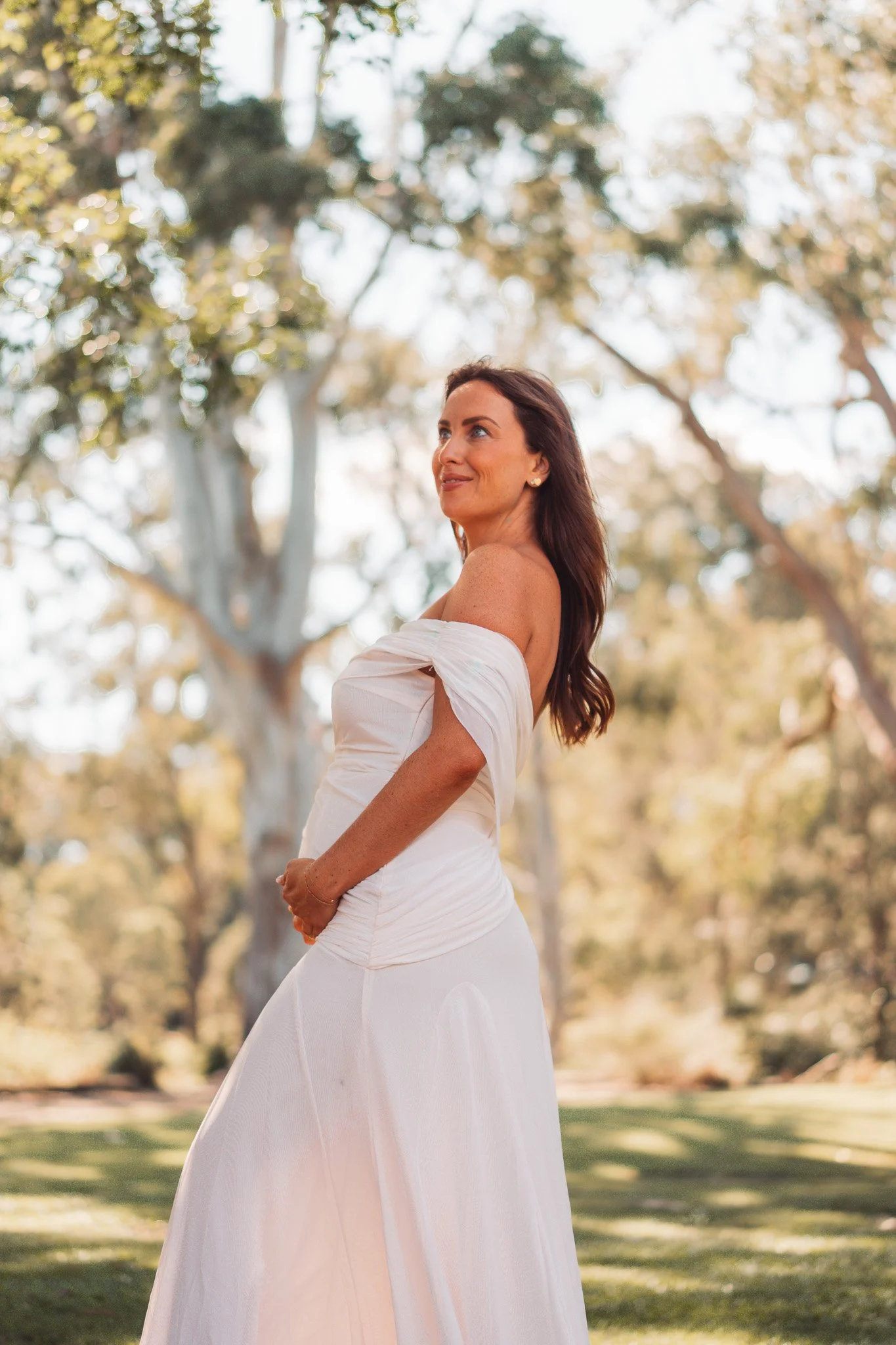 A woman in a white off-shoulder dress standing outdoors among trees with a blurred background, smiling and looking slightly to her left.