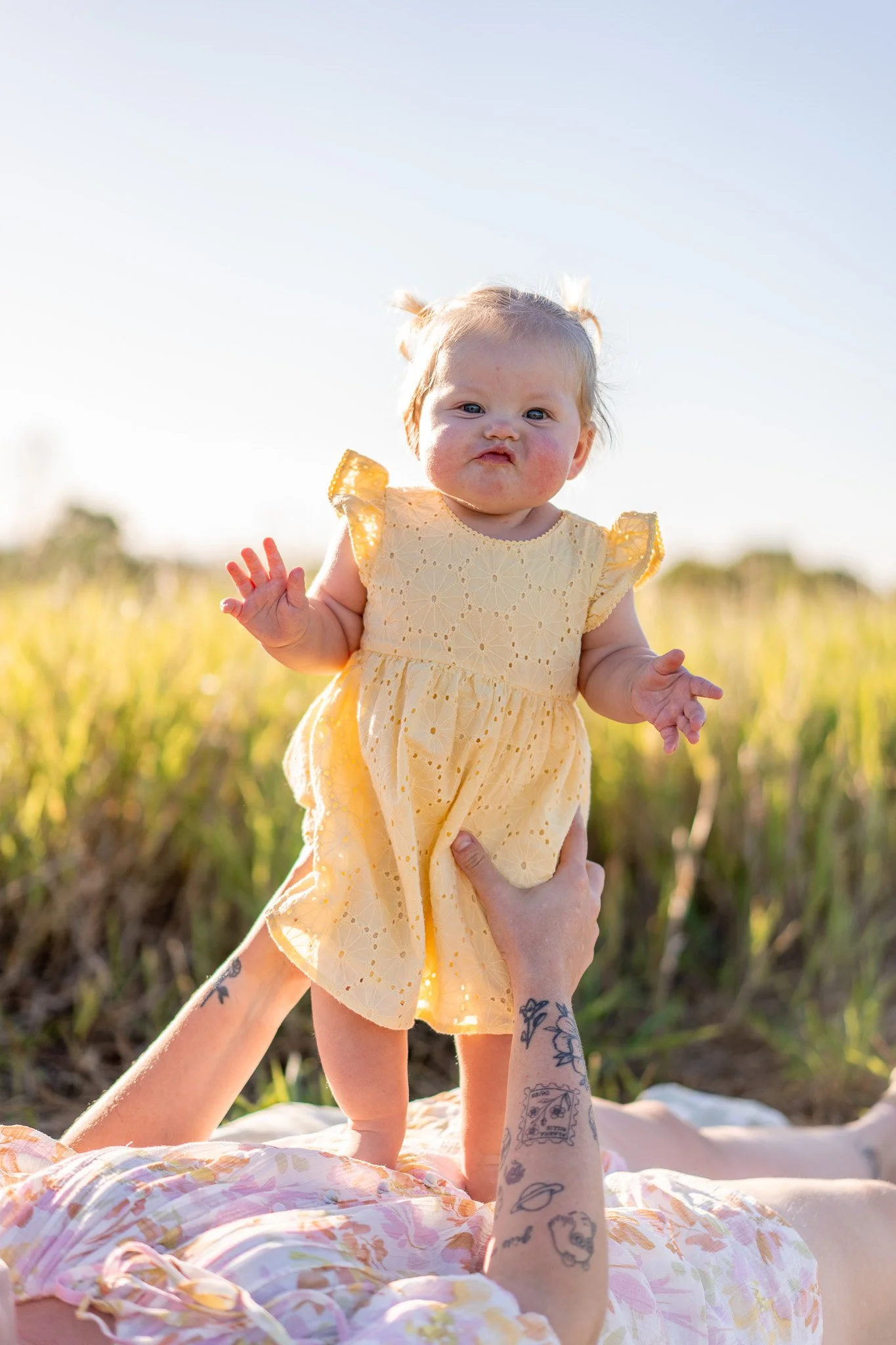 A baby girl in a yellow dress is being held up in the air outdoors during sunset, with a field and blue sky in the background.