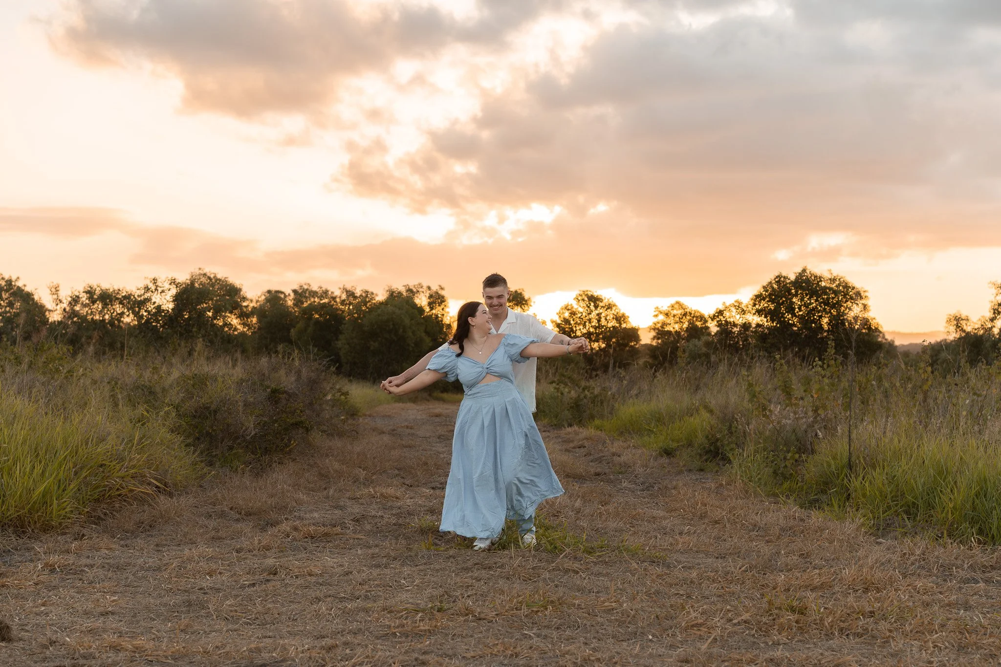 A couple dancing in a field at sunset, with clouds and trees in the background.
