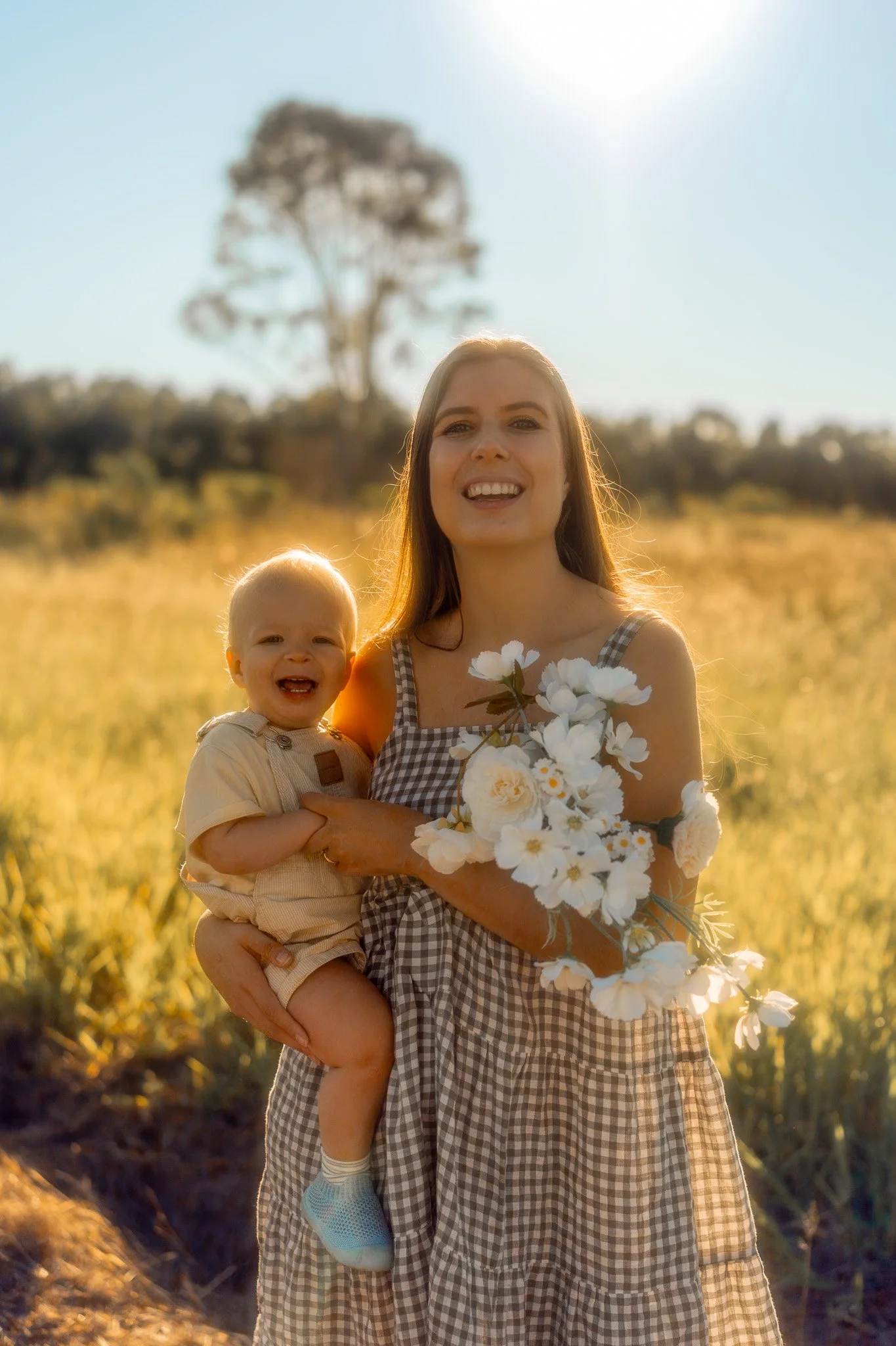 A woman and a young child standing in a field during sunset. The woman is holding a bouquet of white flowers and smiling, while the child is also smiling and looking at the camera. There is a large tree in the background and the sky is clear with the sun shining brightly.