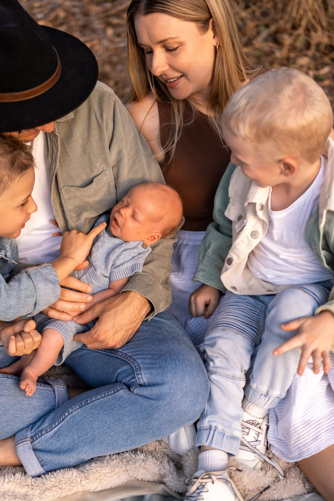 A woman with blonde hair smiling and looking at a newborn baby being held by a man wearing a hat. Two young children, one with blonde hair and the other with brown hair, are also looking at the baby. They are outdoors, on a fuzzy rug, with a backgrou