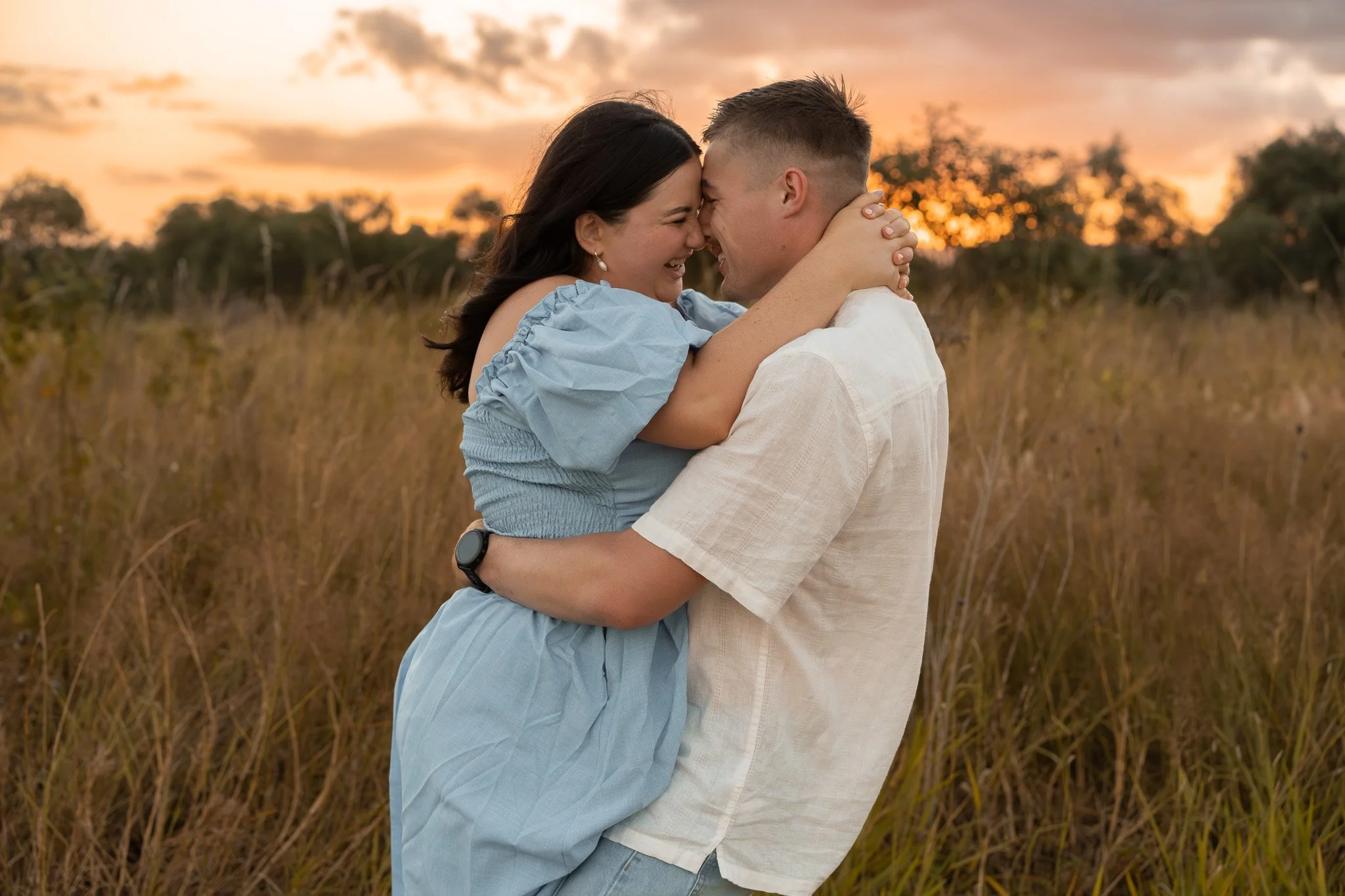A couple in a field at sunset, embracing and touching foreheads, smiling with joy.