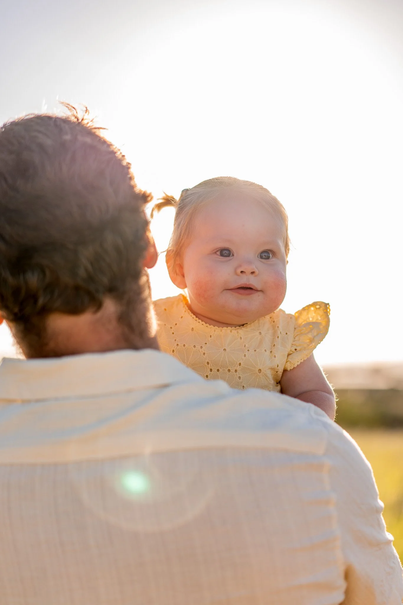 A baby girl with light skin and red hair being held by an adult during sunset, with bright sunlight in the background.