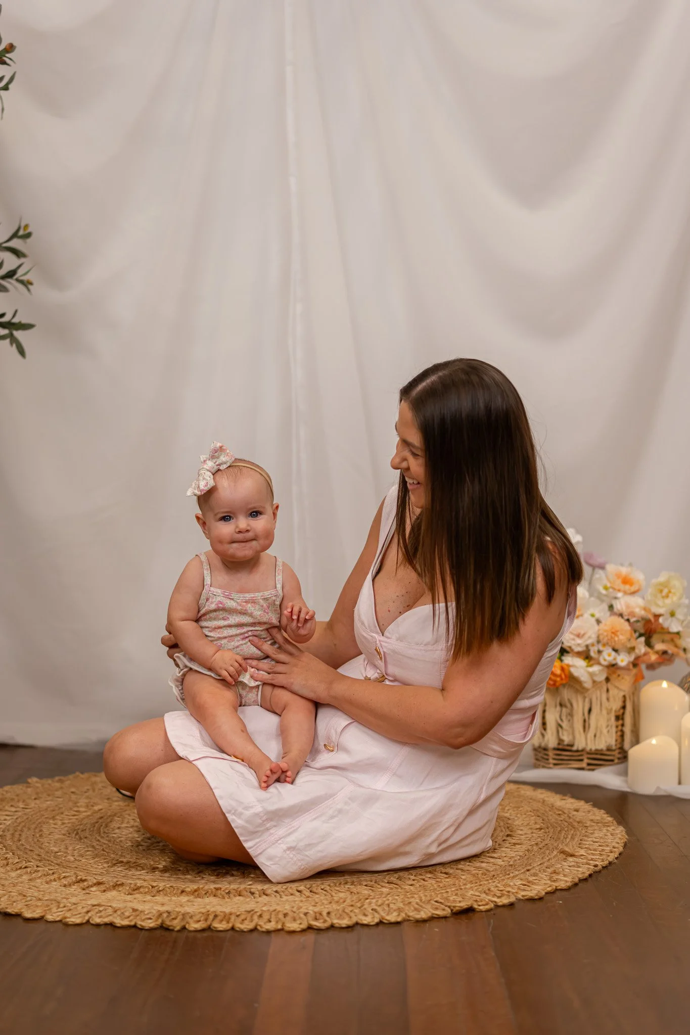 A woman sitting cross-legged on a circular rug holding a toddler girl. The girl has a floral bow headband and is wearing a floral dress. The woman is smiling and looking at the girl, who has a neutral expression. Behind them is a white curtain, with 